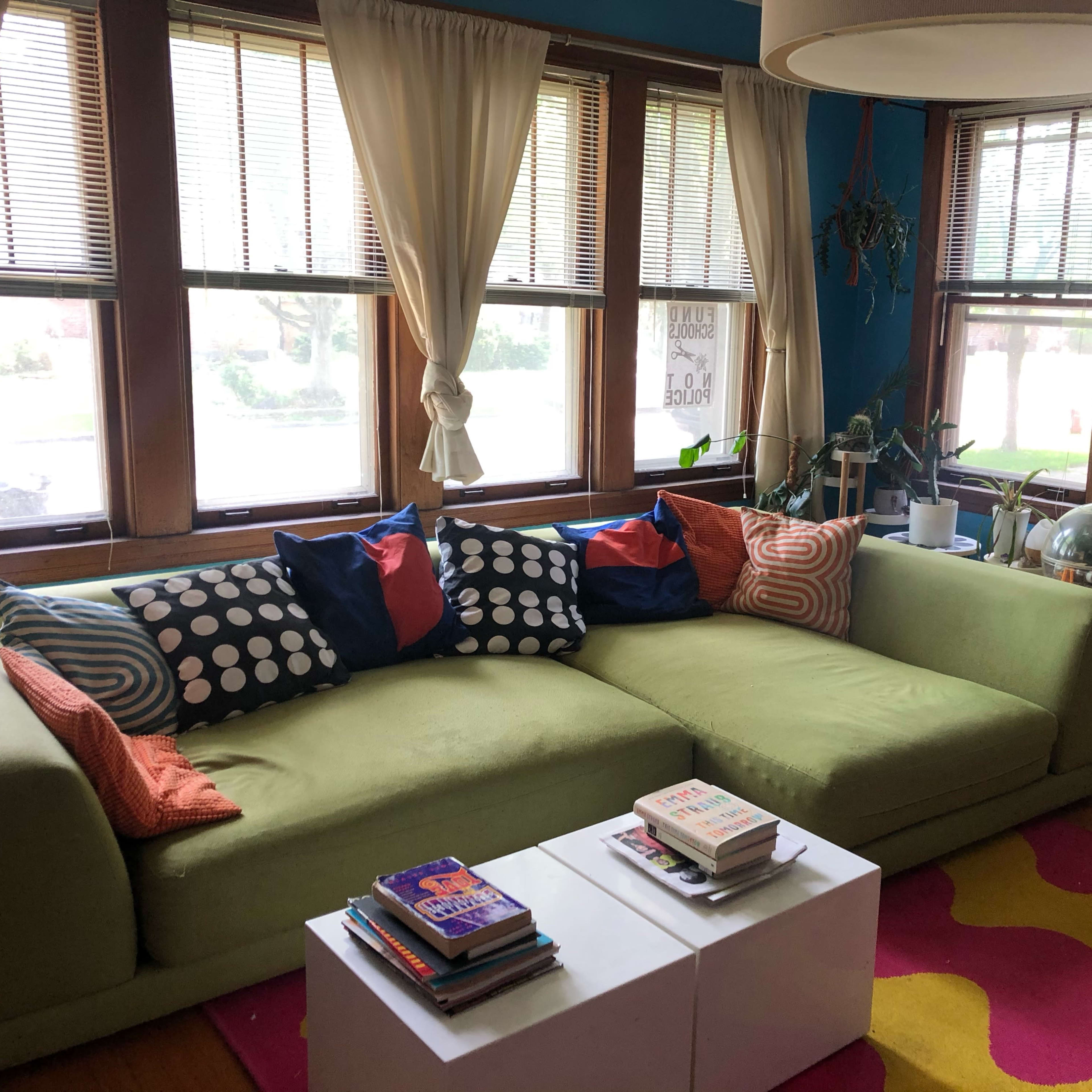 A green sectional sofa with colorful throw pillows is positioned against a wall of windows, accompanied by a coffee table and a collection of books on the floor in a brightly lit room.