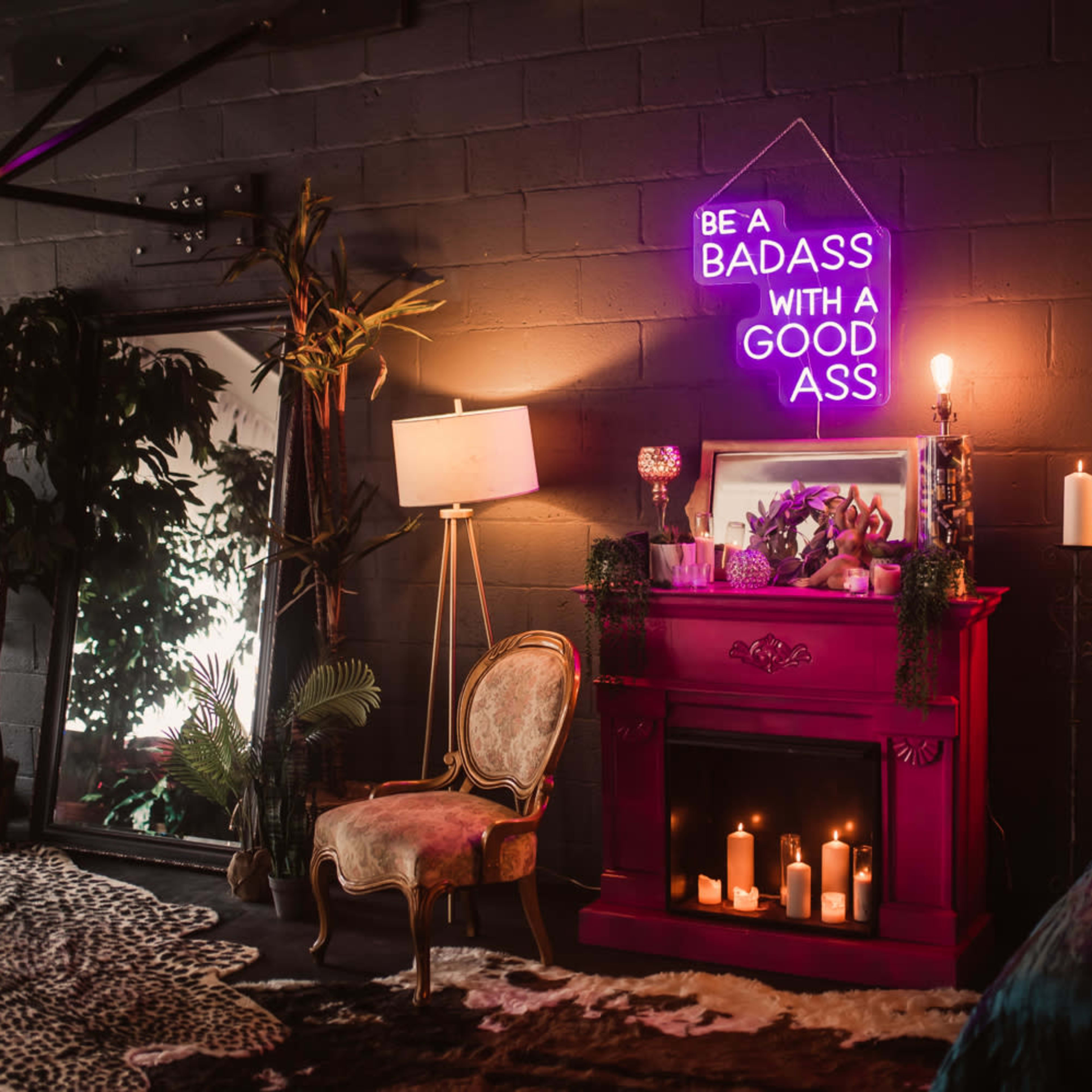 A dimly lit room featuring a bright pink fireplace adorned with candles, a neon sign, a vintage chair, and decorative plants.