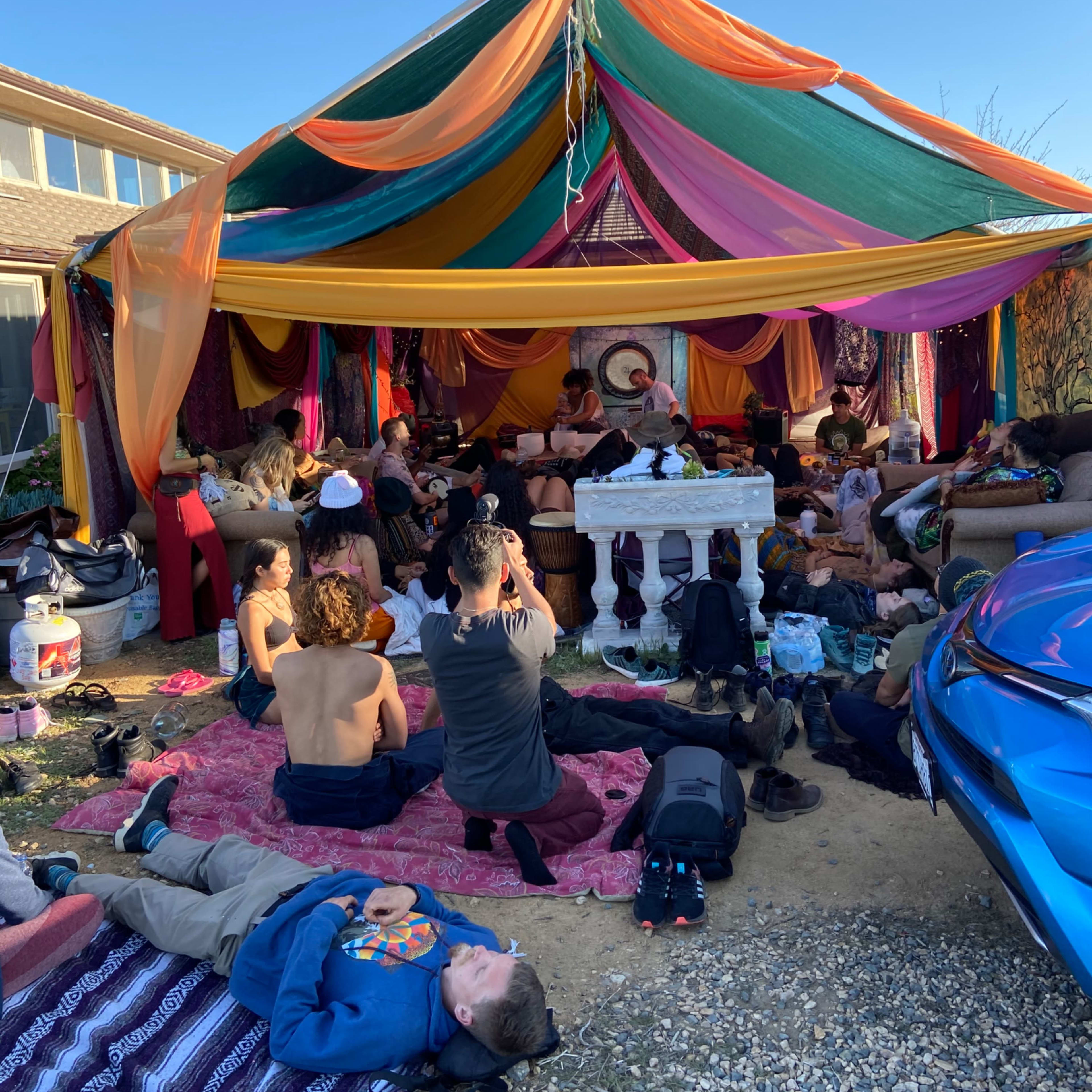 A large colorful canopy covers a gathering of people sitting on the ground and lounging in a casual outdoor setting.