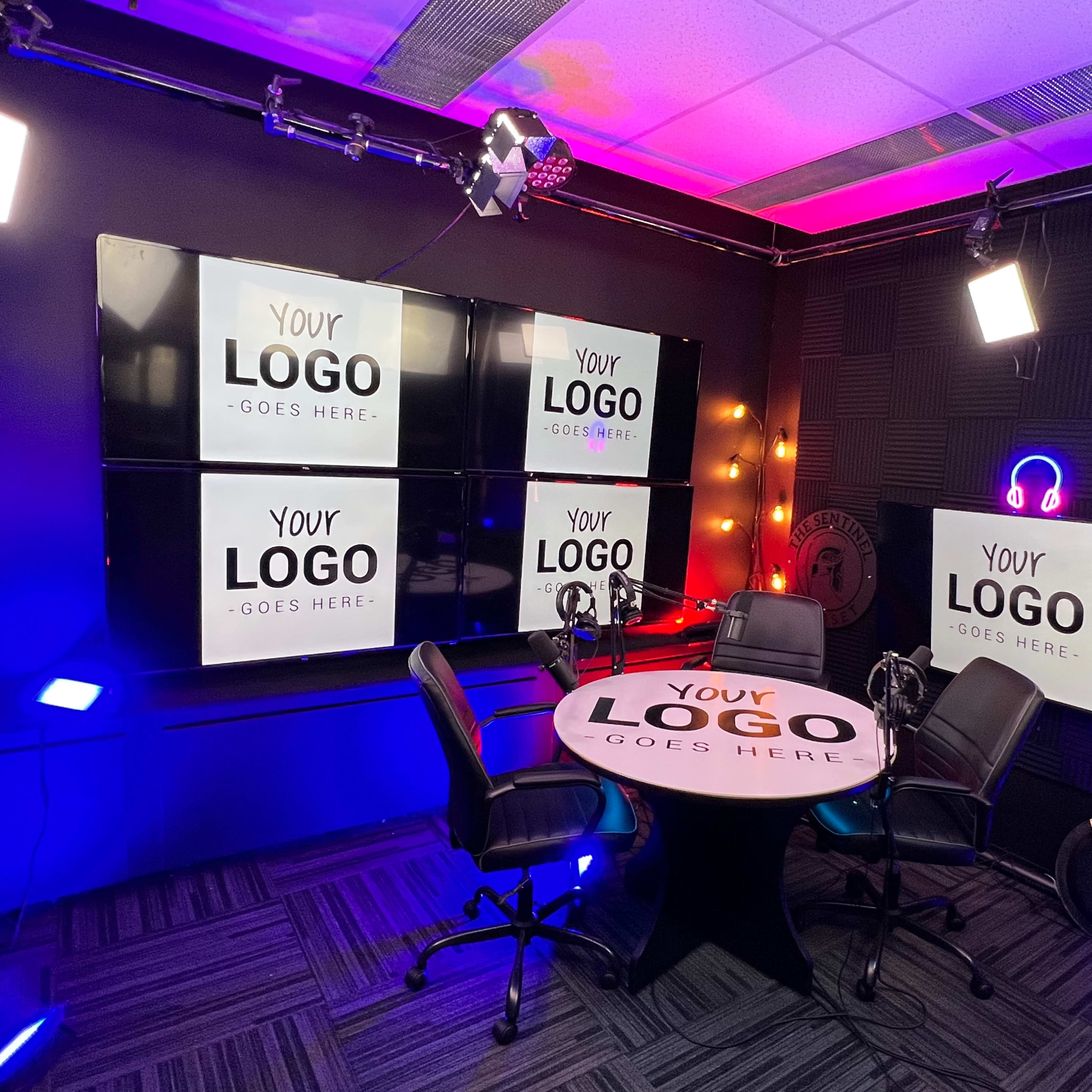 A modern studio setup for video production, featuring a round table with two chairs, multiple large screens displaying the text "YOUR LOGO GOES HERE," and various lighting fixtures.