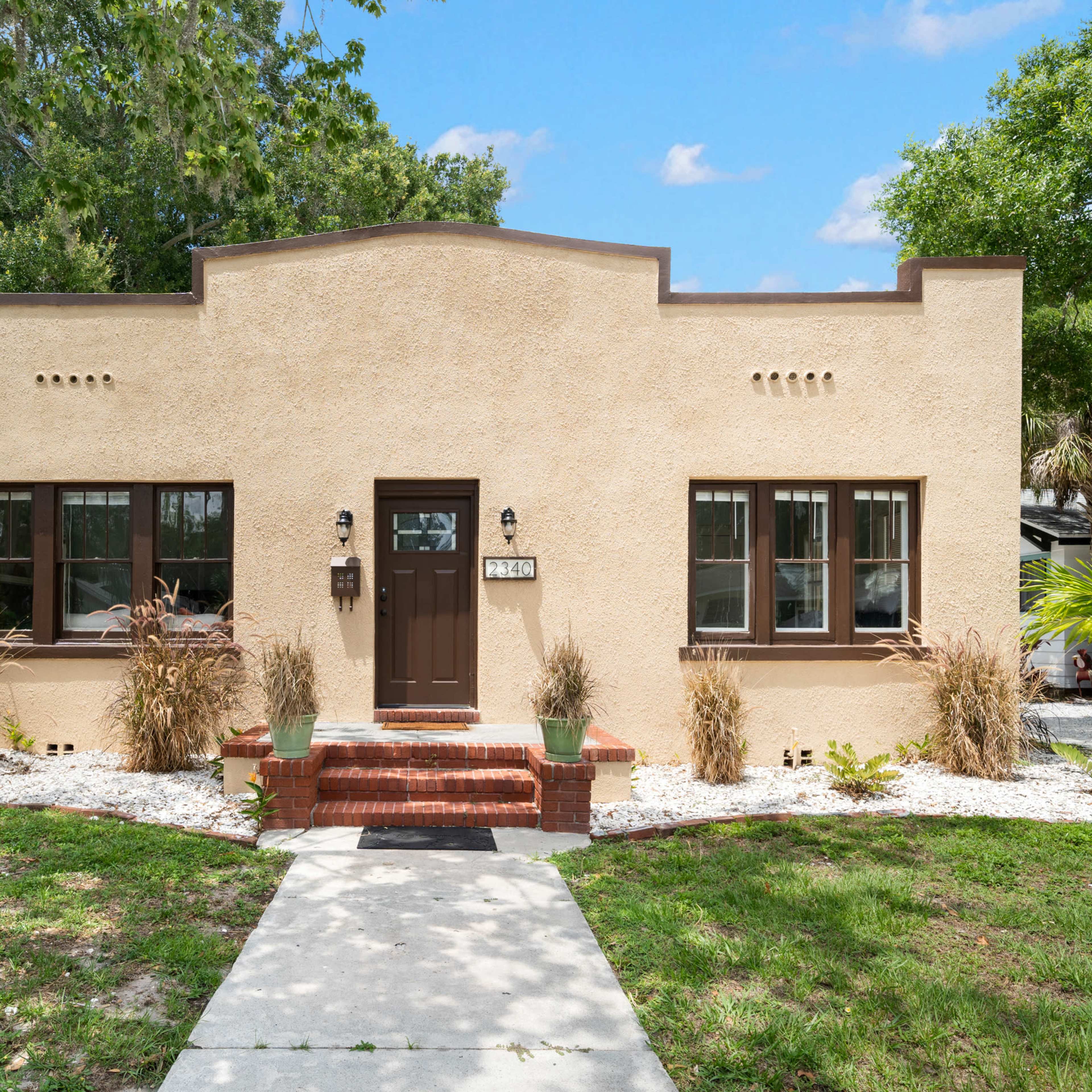 A single-story, stucco house with a brown door and windows, surrounded by landscaped grass and decorative plants.