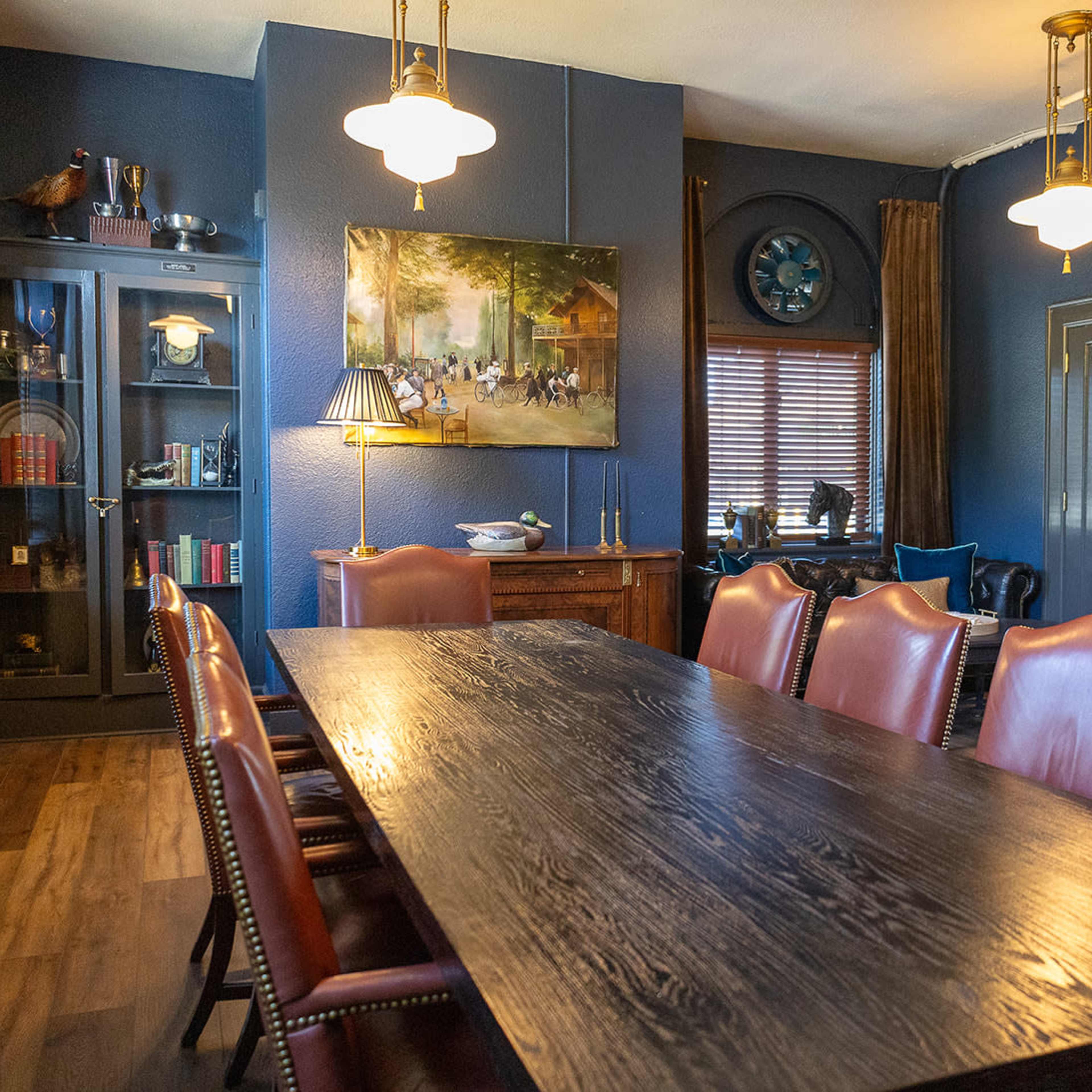 A dining room with a long wooden table surrounded by leather chairs, accented by a large painting and shelves filled with decorative items.