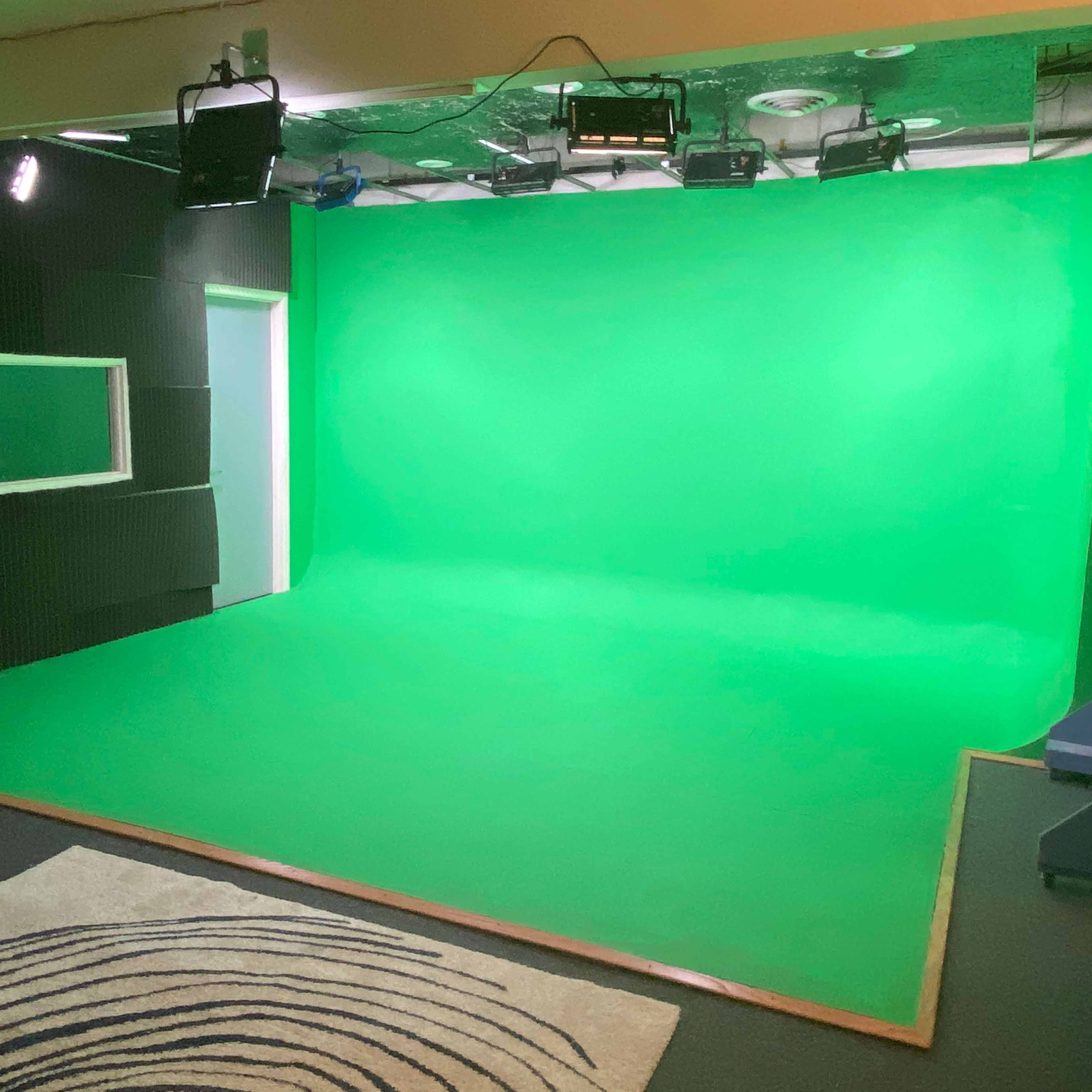 A green screen studio with a flat green backdrop, track lighting, and a small window in the black acoustic wall.