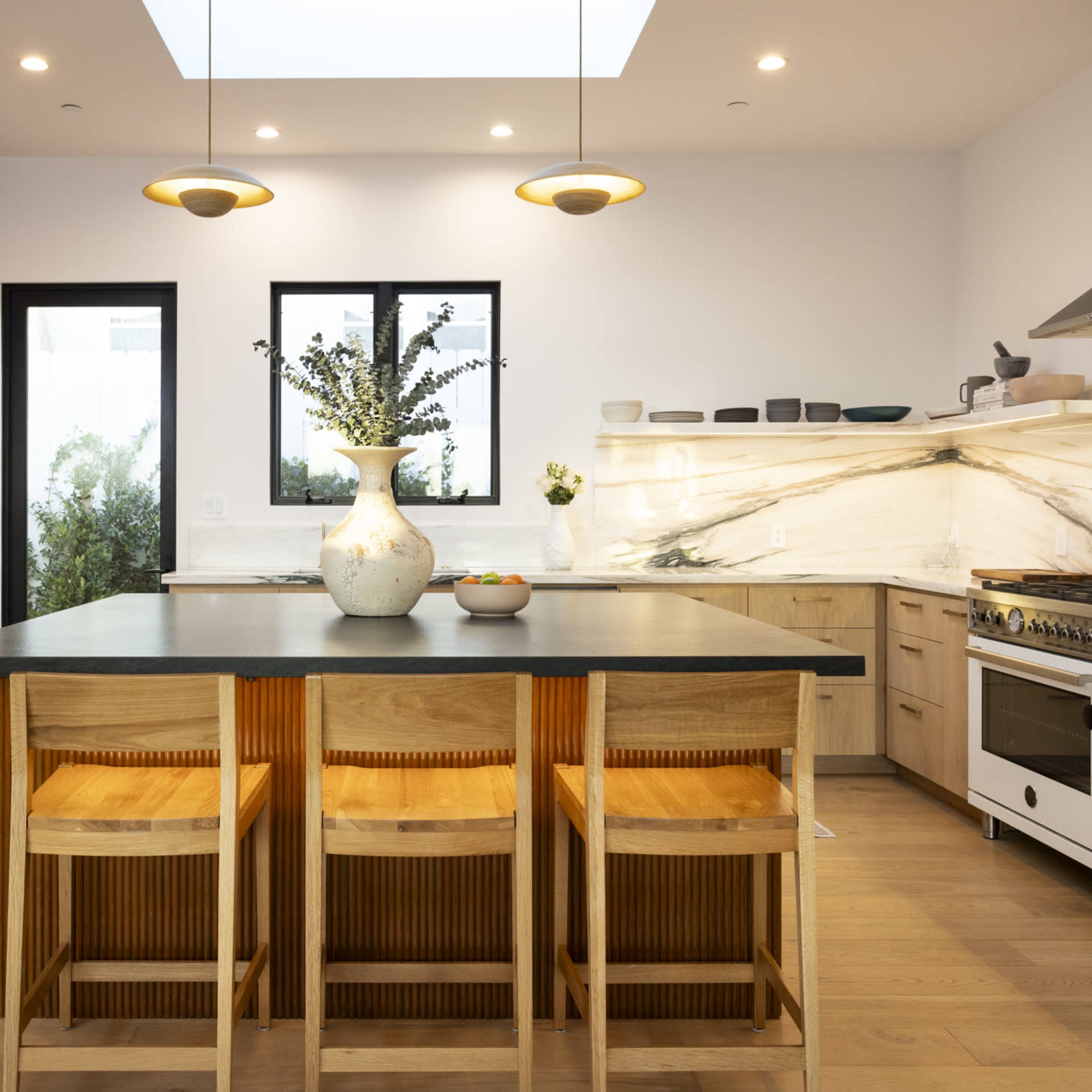 A modern kitchen features light wood cabinetry, a central island with bar stools, and a marble backsplash with a skylight above.