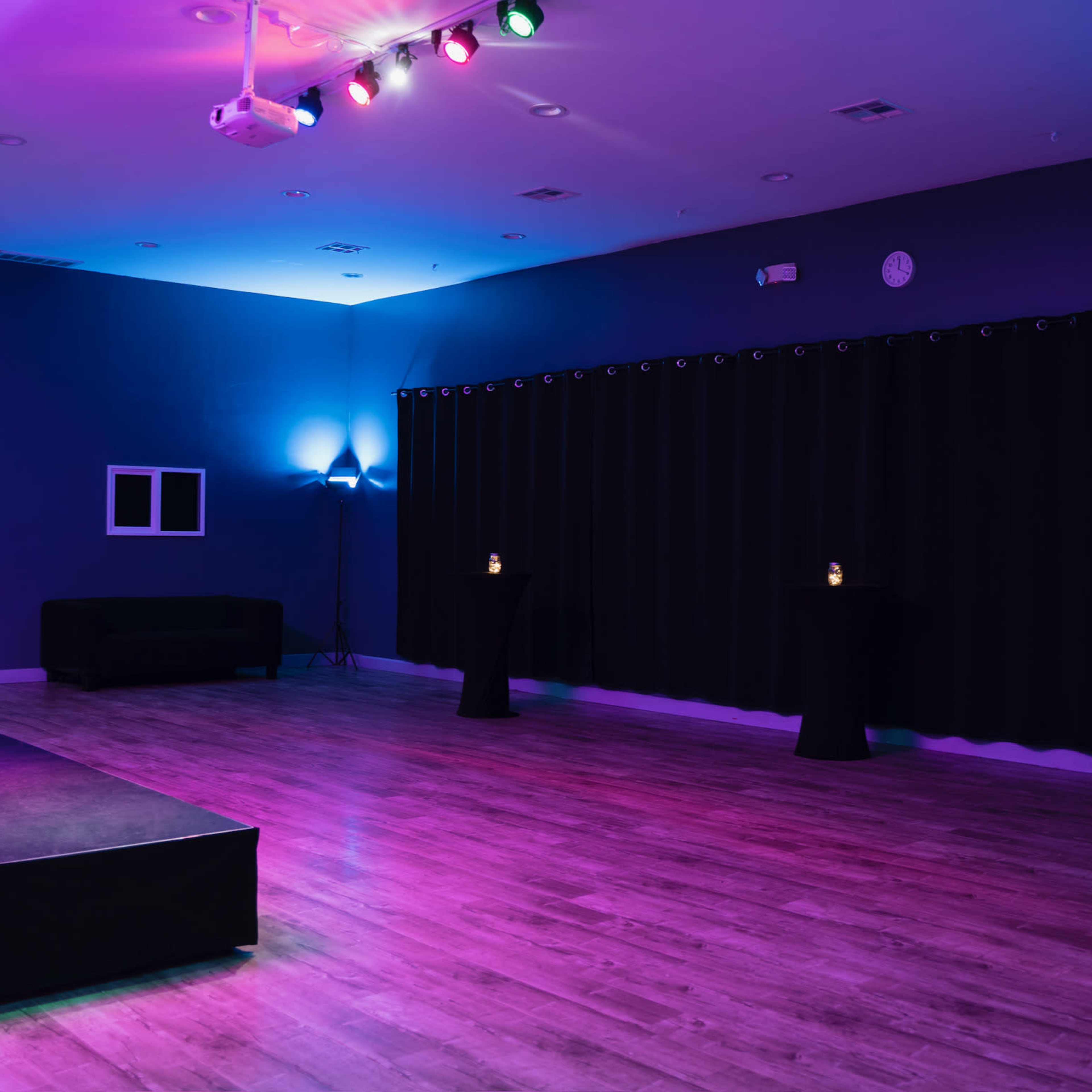 A dimly lit event space features a small stage, black curtains, and several illuminated pedestal tables.