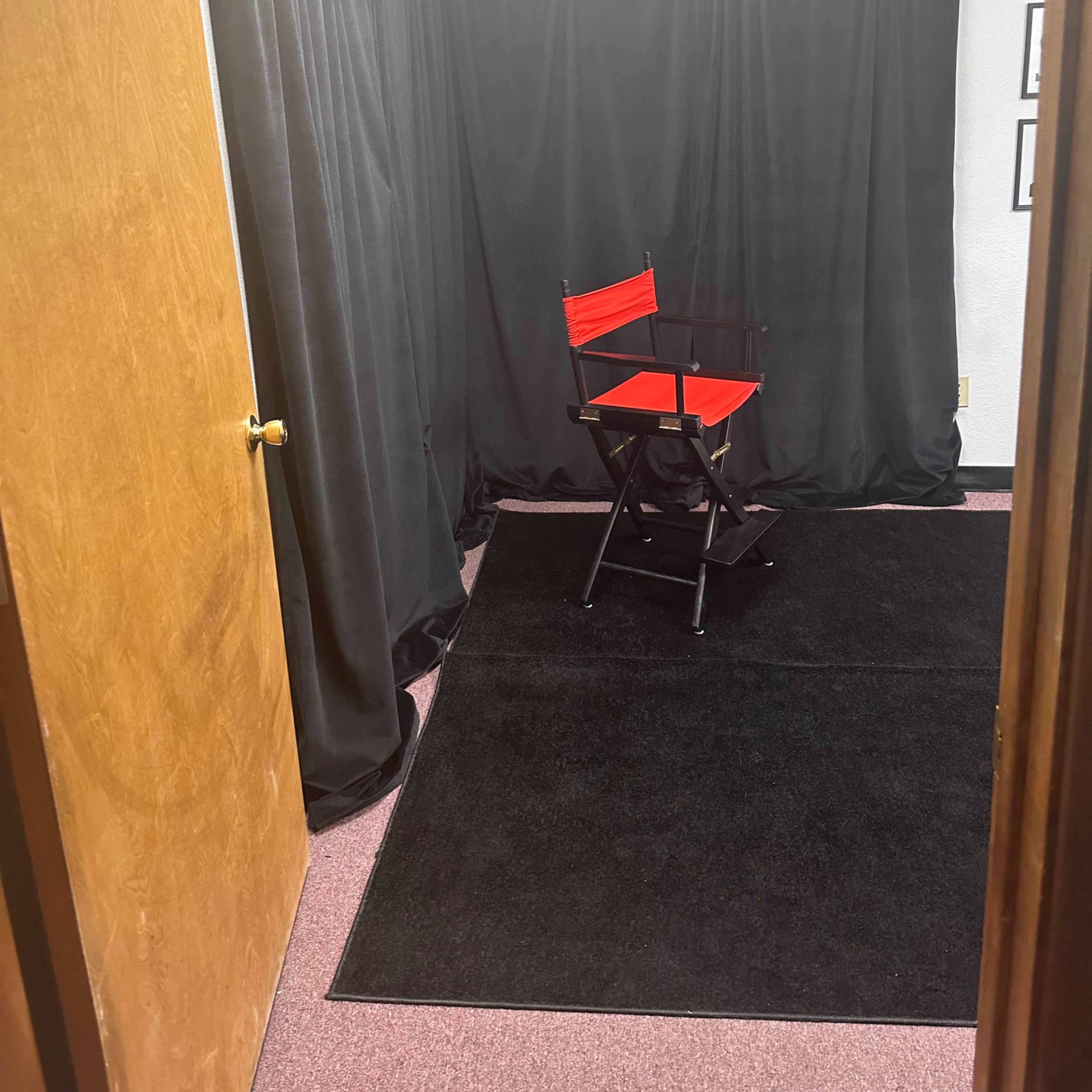 A small room with a black curtain backdrop, a red director's chair, and a dark carpet, with a wooden door partially open.