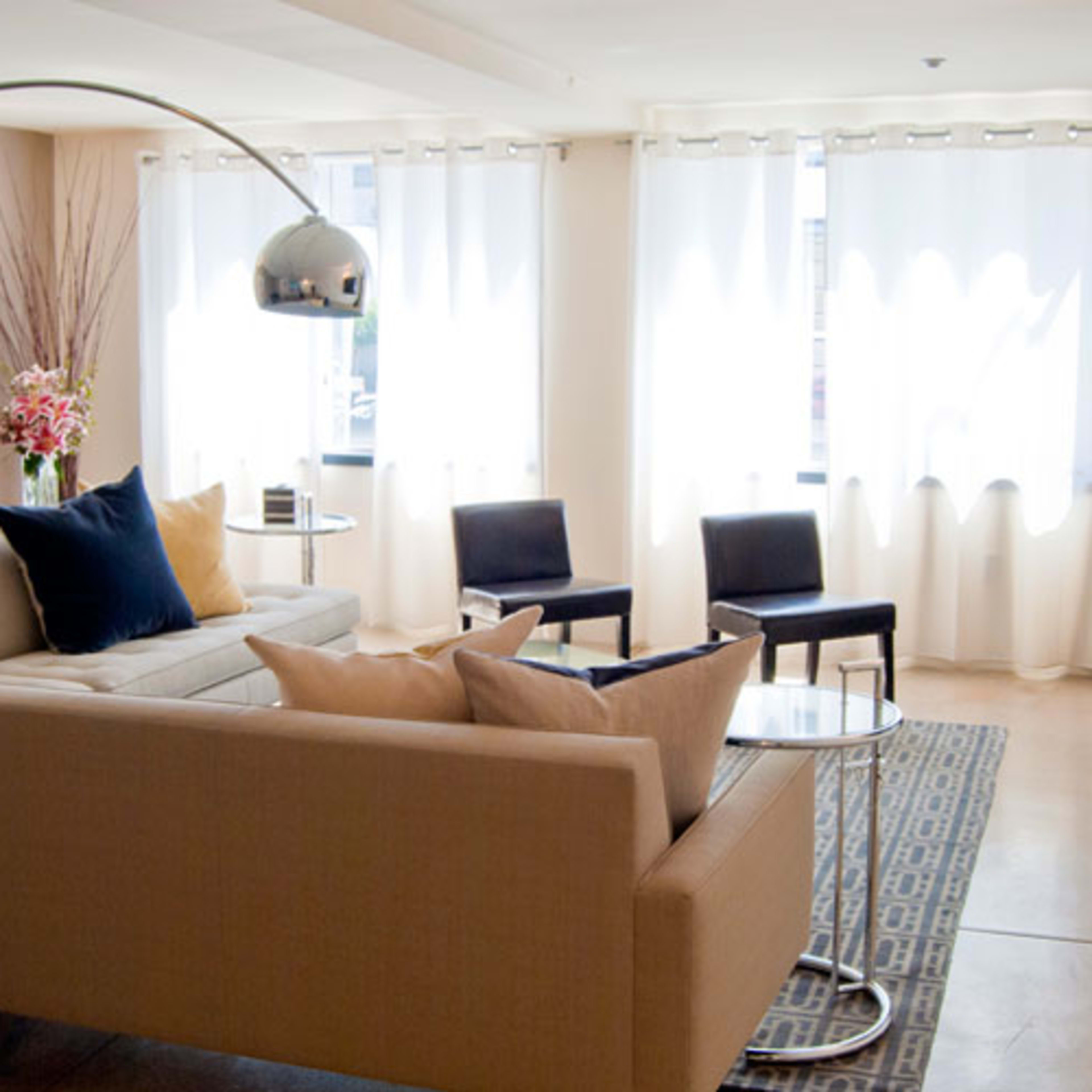 A modern living room with a curved sofa, a glass coffee table, and several chairs, illuminated by natural light through sheer curtains.