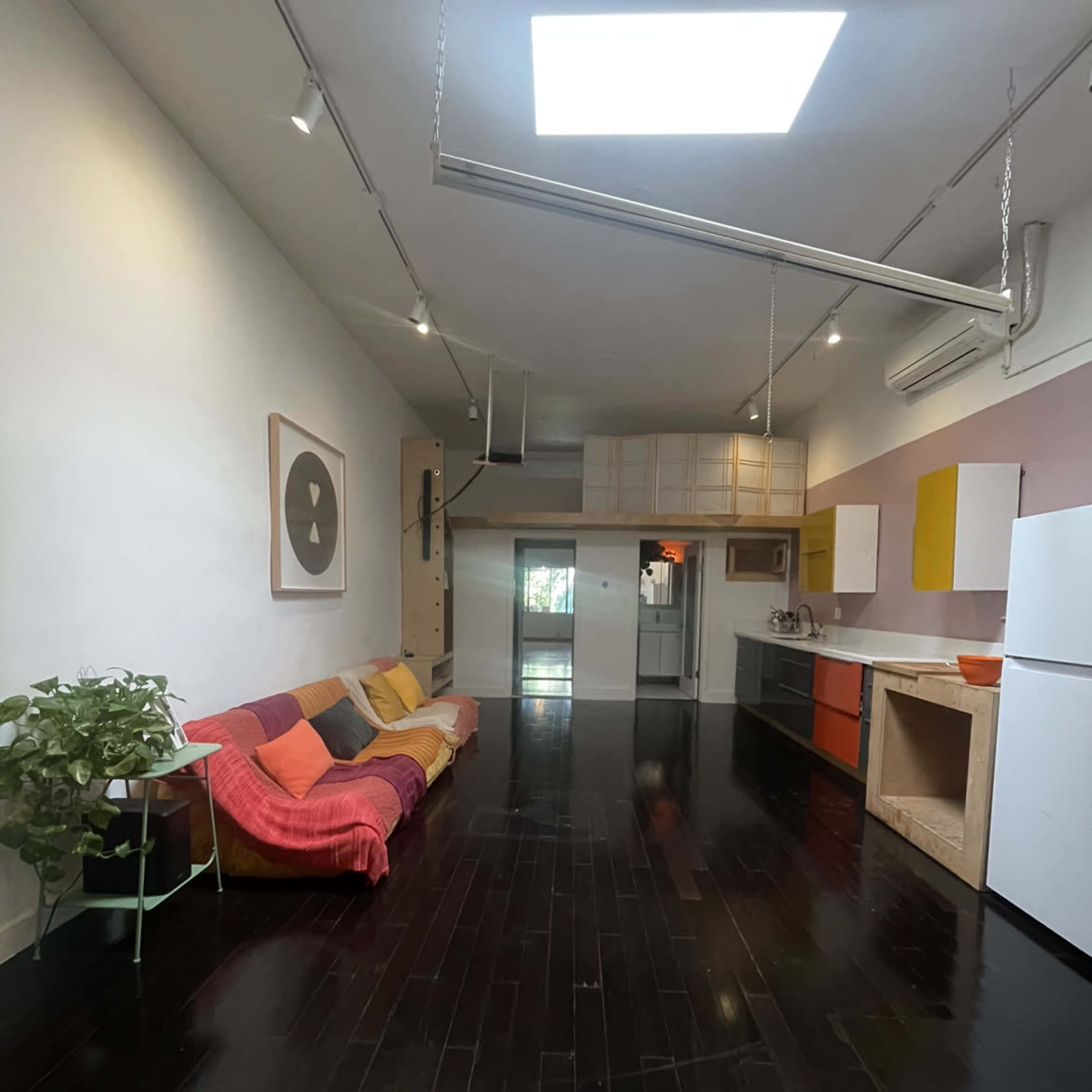A spacious interior features a colorful sectional sofa, a minimalist kitchen with bright cabinetry, and large windows allowing natural light.