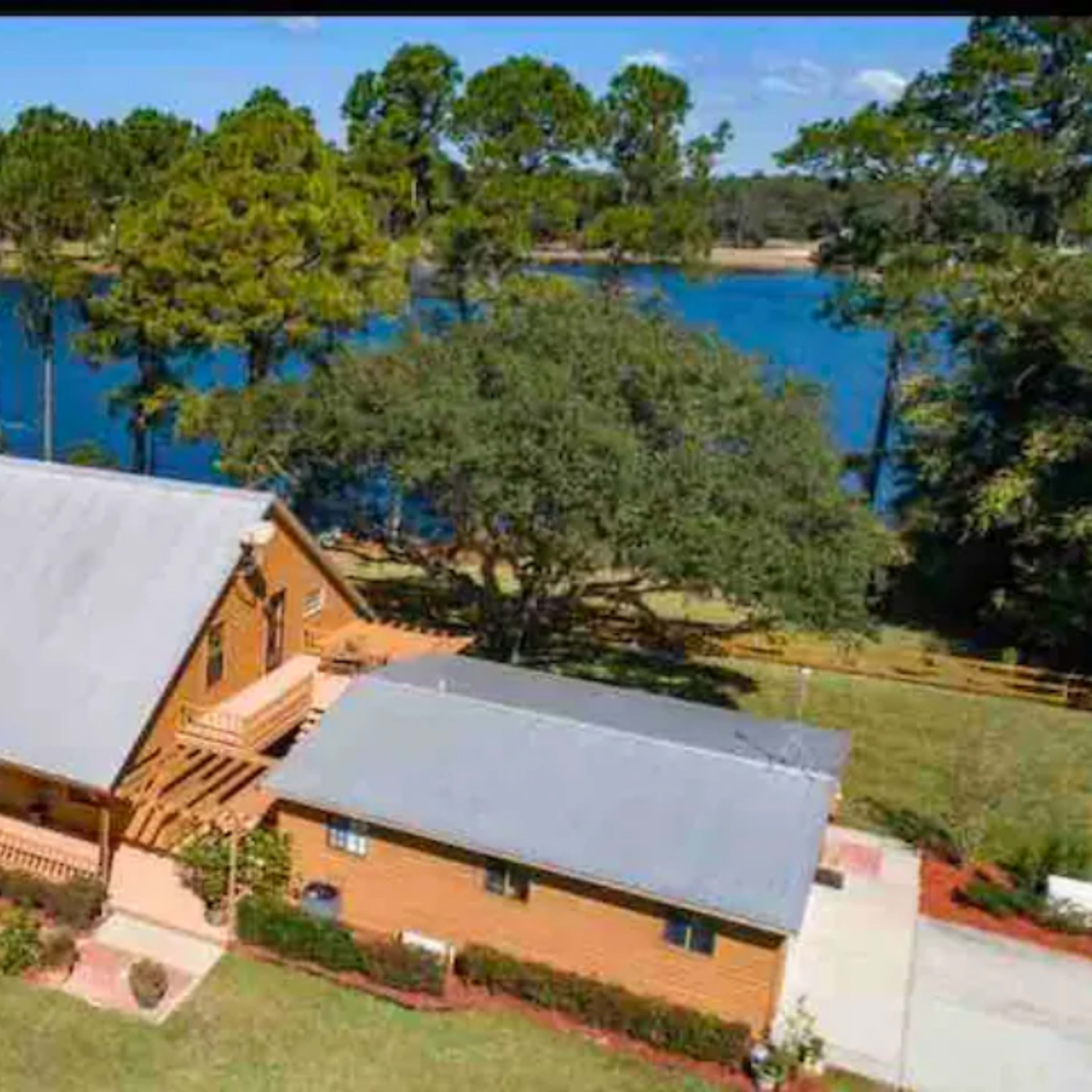 A two-story wooden house beside a lake, surrounded by trees and greenery.