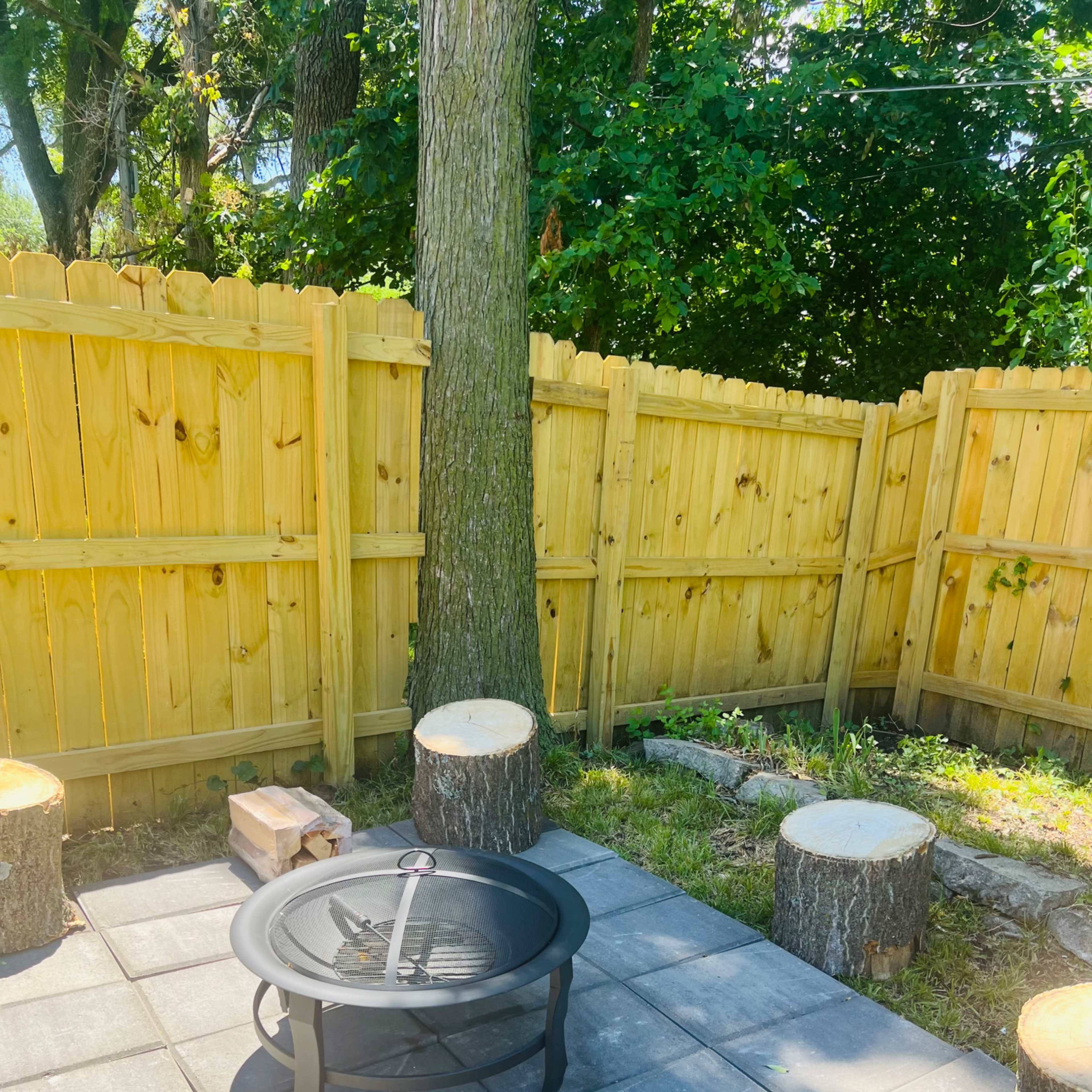 A fire pit surrounded by stone slabs and tree stump seating, enclosed by a wooden fence and greenery.