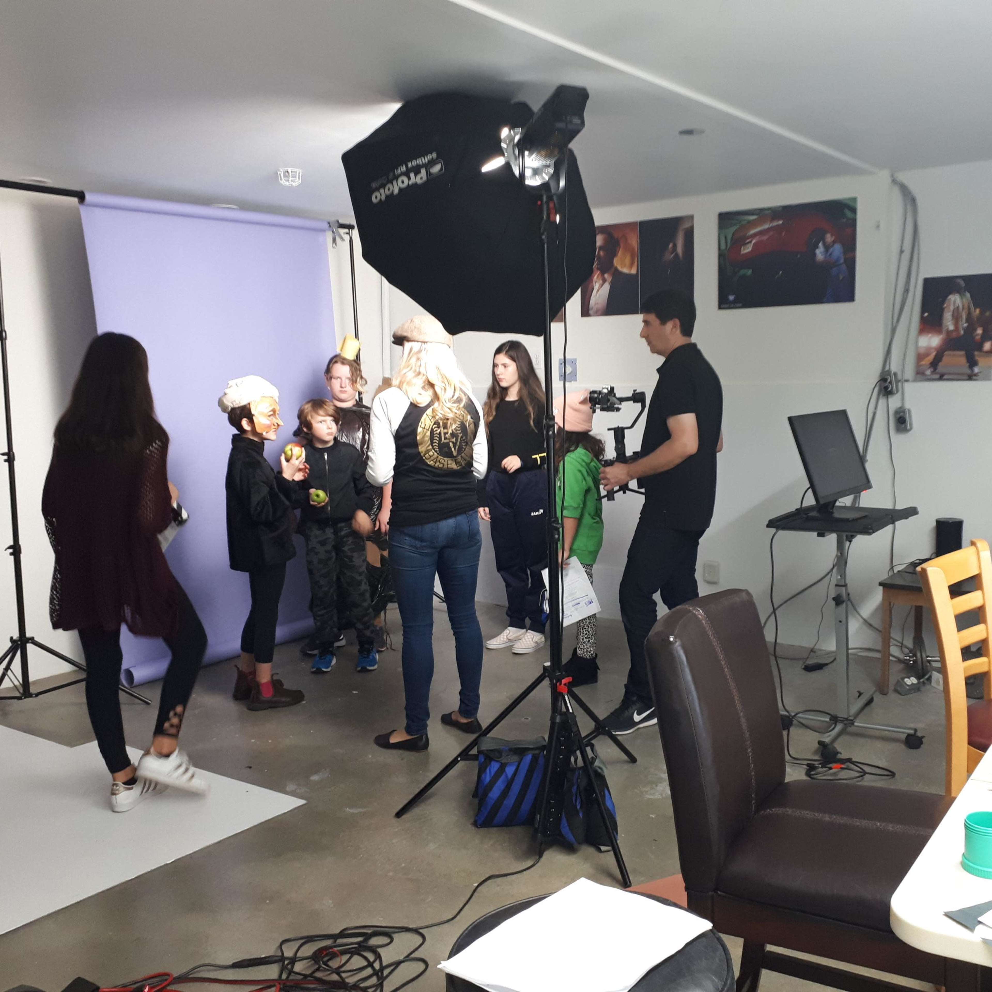 A group of children and adults are gathered in a photography studio, preparing for a photoshoot with various lighting equipment and backdrops set up.