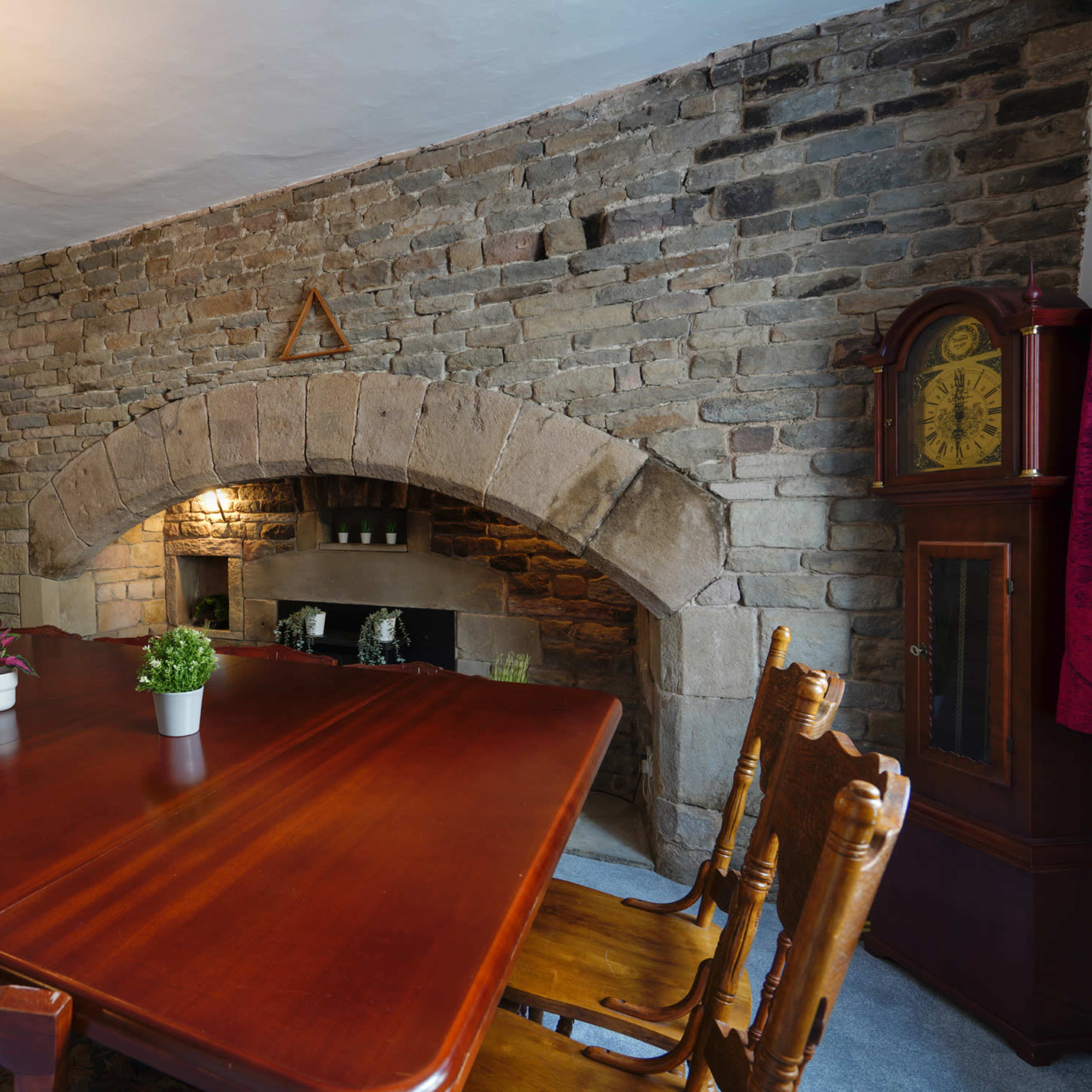 A dining room with a large wooden table, surrounded by high-backed chairs, and a stone fireplace set into a rough stone wall.
