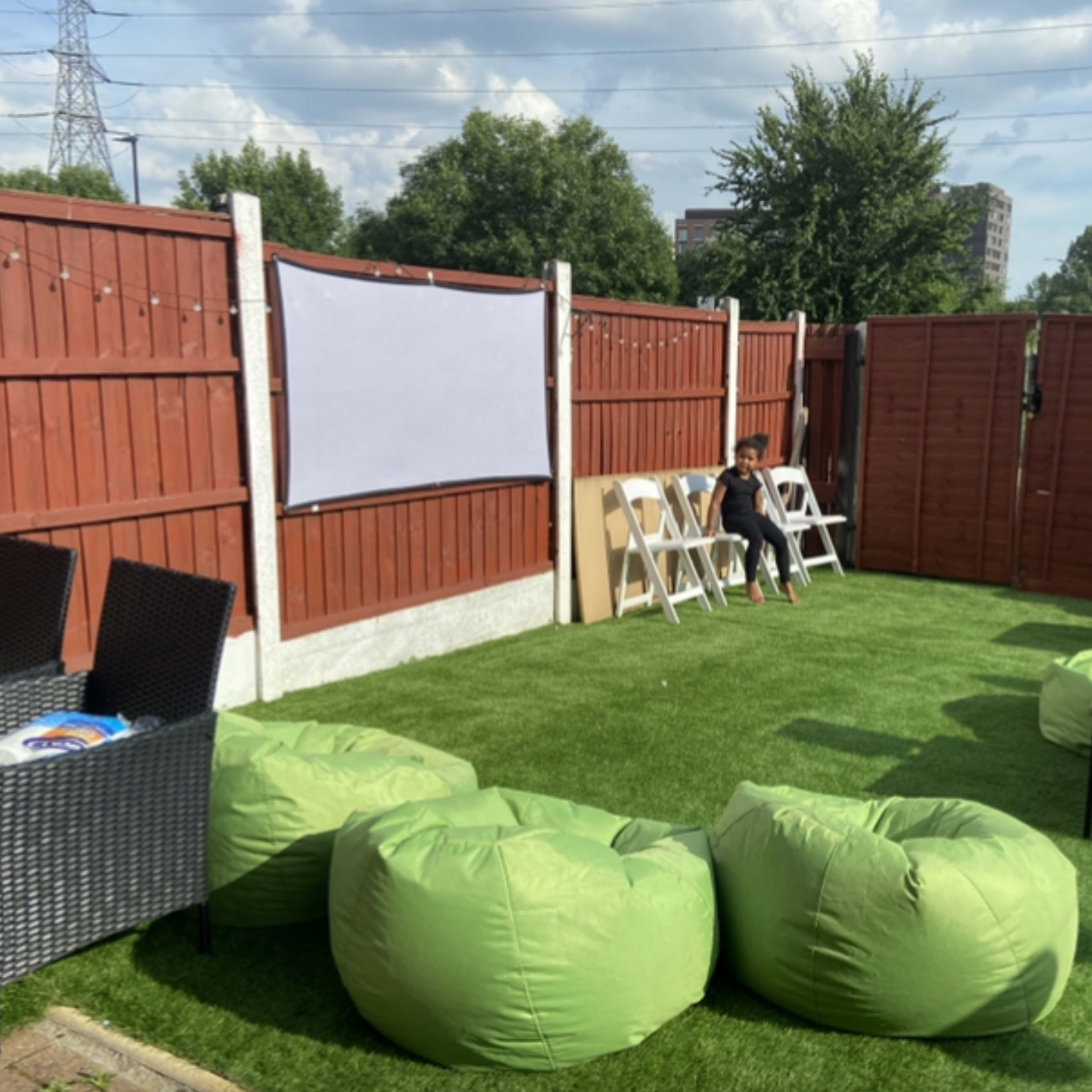 A person sits on a chair in a fenced backyard decorated with green bean bag chairs and a white projector screen.