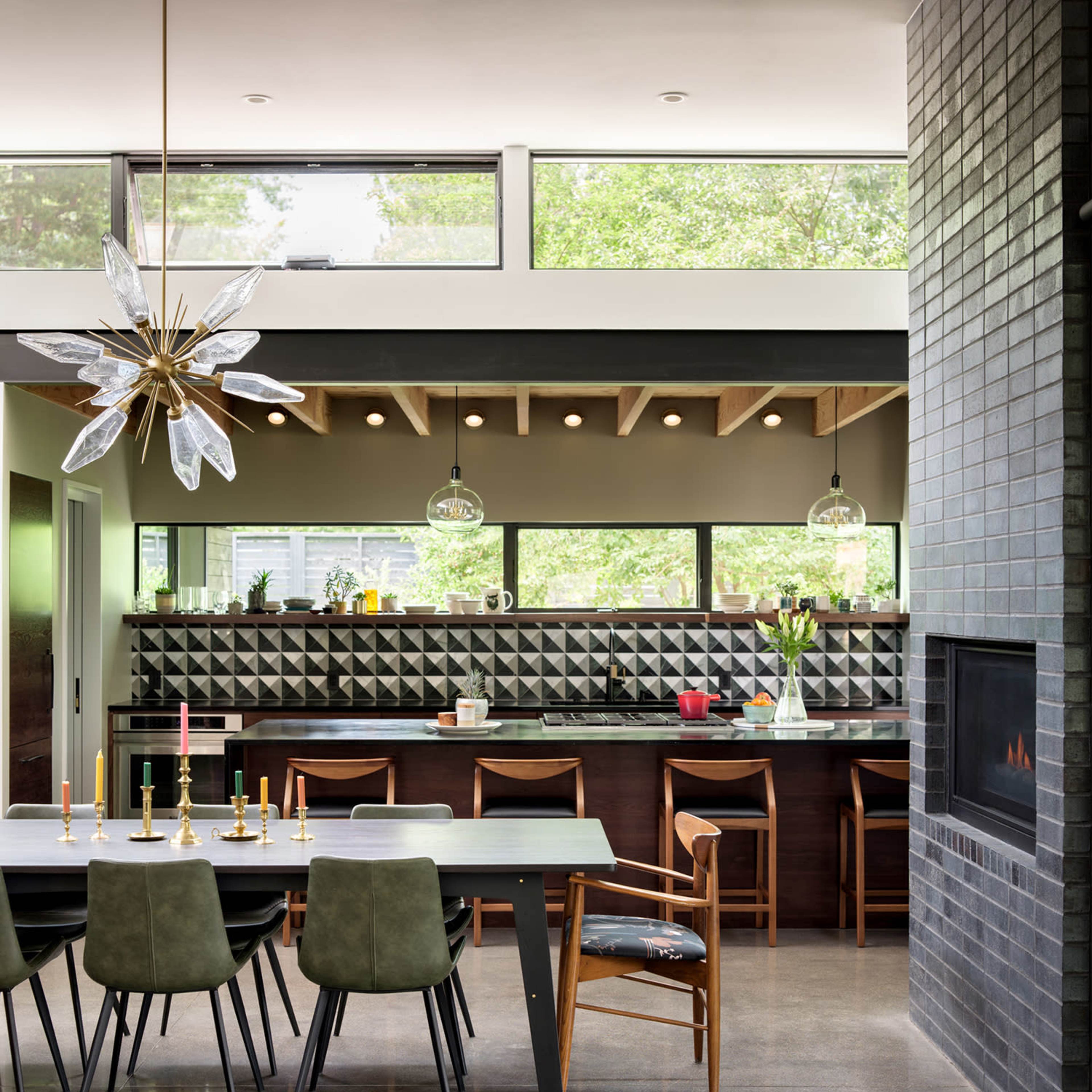 A modern kitchen and dining area features a long table with chairs, a sleek counter with geometric tile backsplash, and large windows letting in natural light.