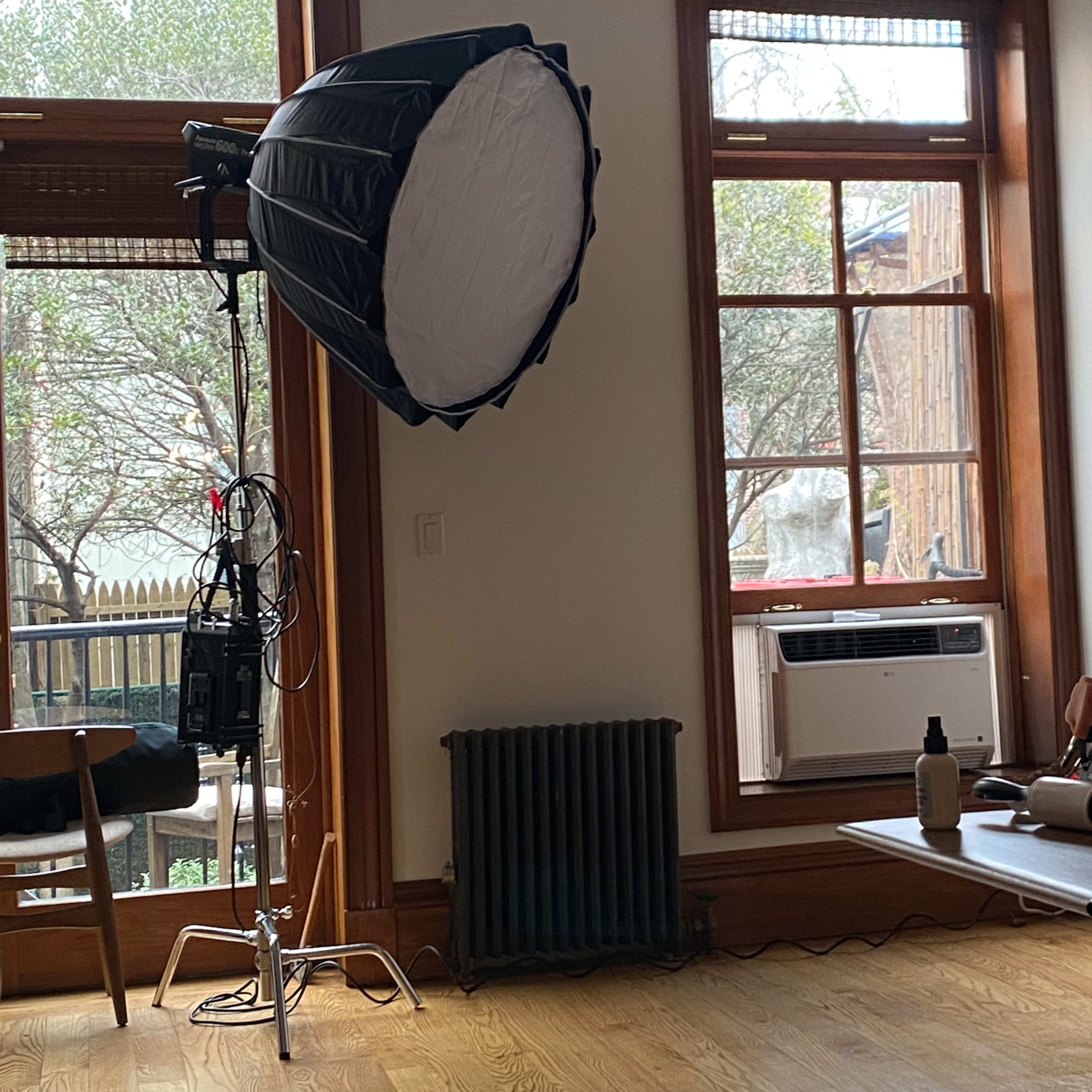 A well-lit room with a large photography light setup beside a window, an air conditioning unit, and various photography equipment on a table.