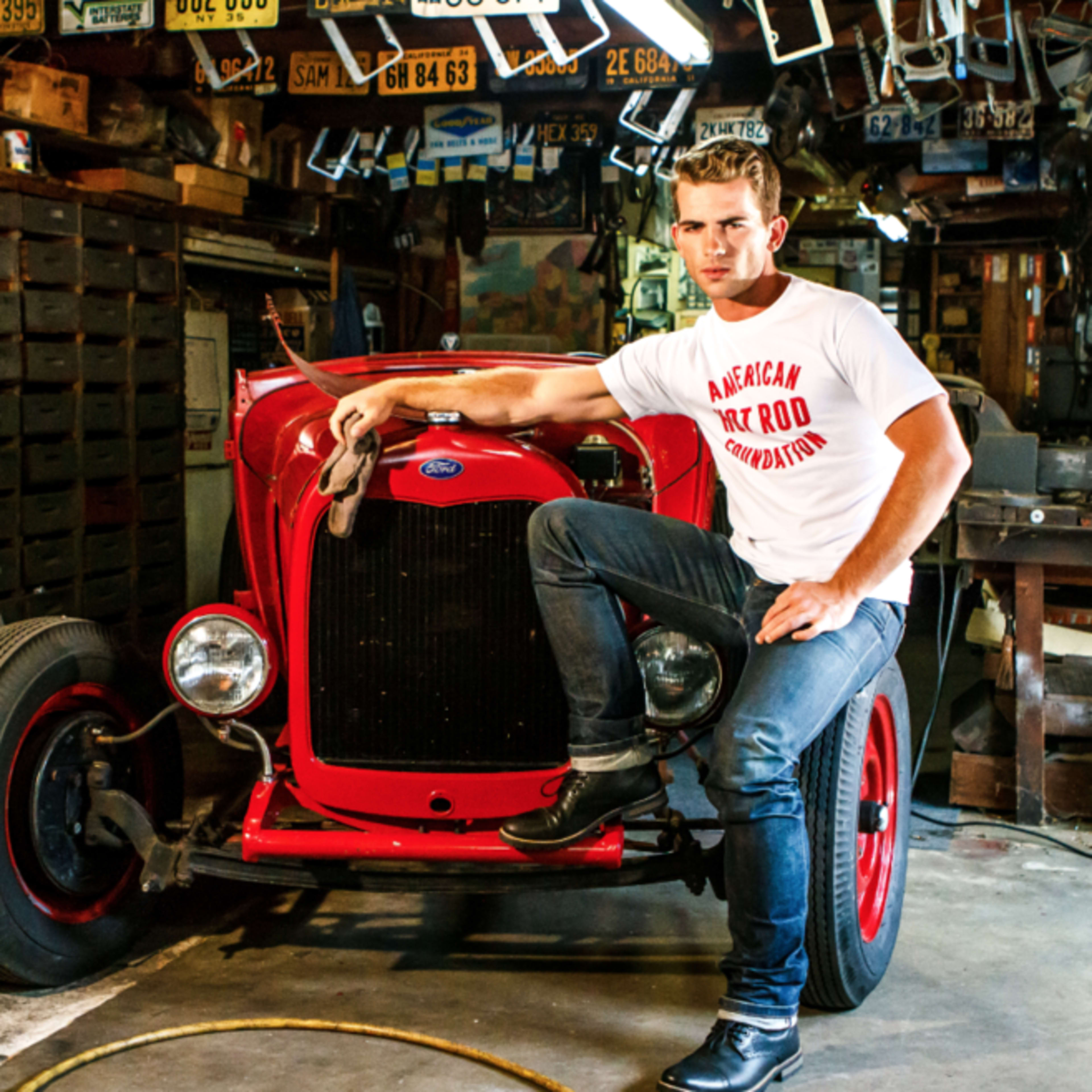 A man in a white t-shirt leans against a red vintage car in a cluttered garage filled with automotive tools and memorabilia.