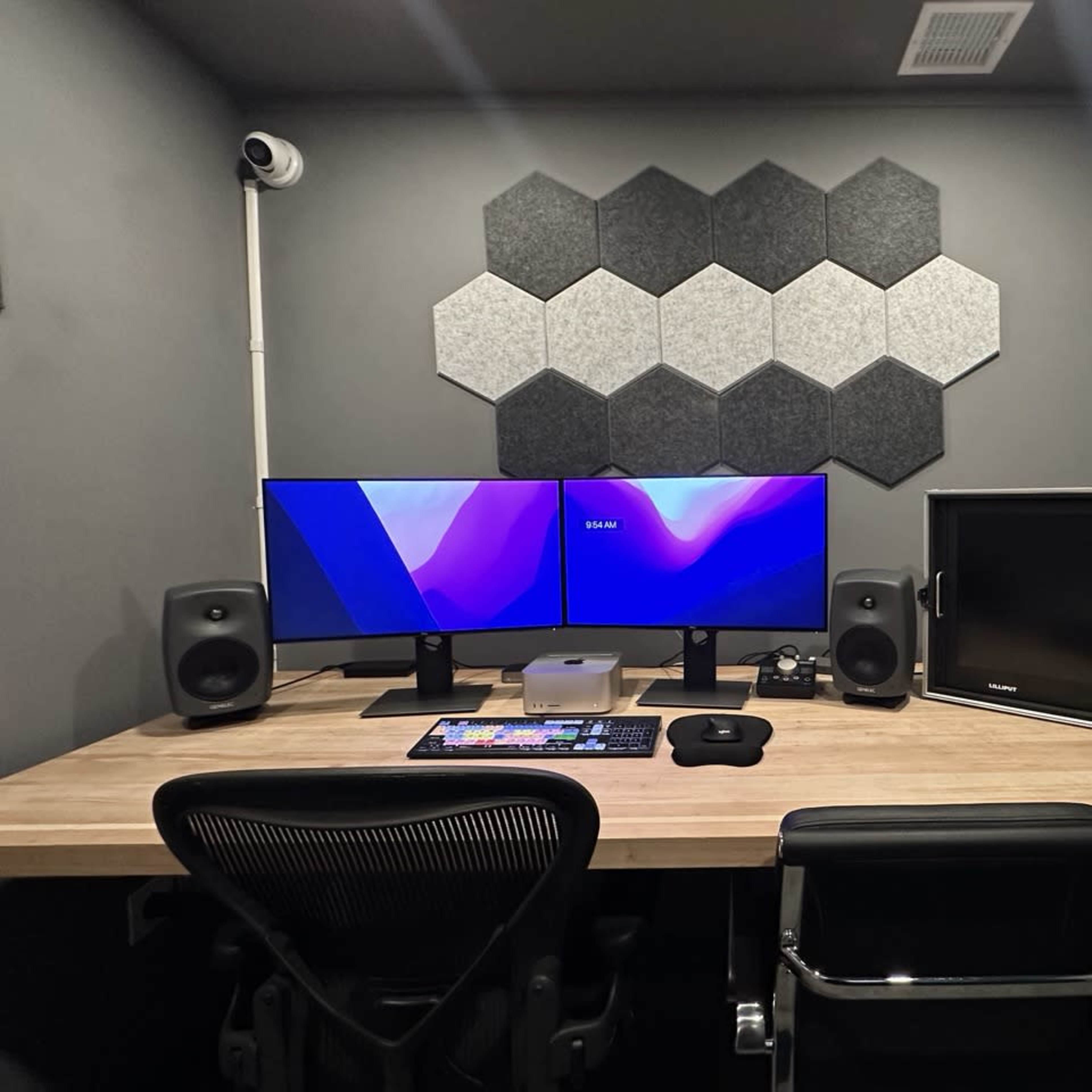 A modern workspace featuring a wooden desk with two monitors, speakers, and a keyboard, against a wall adorned with hexagonal acoustic panels.