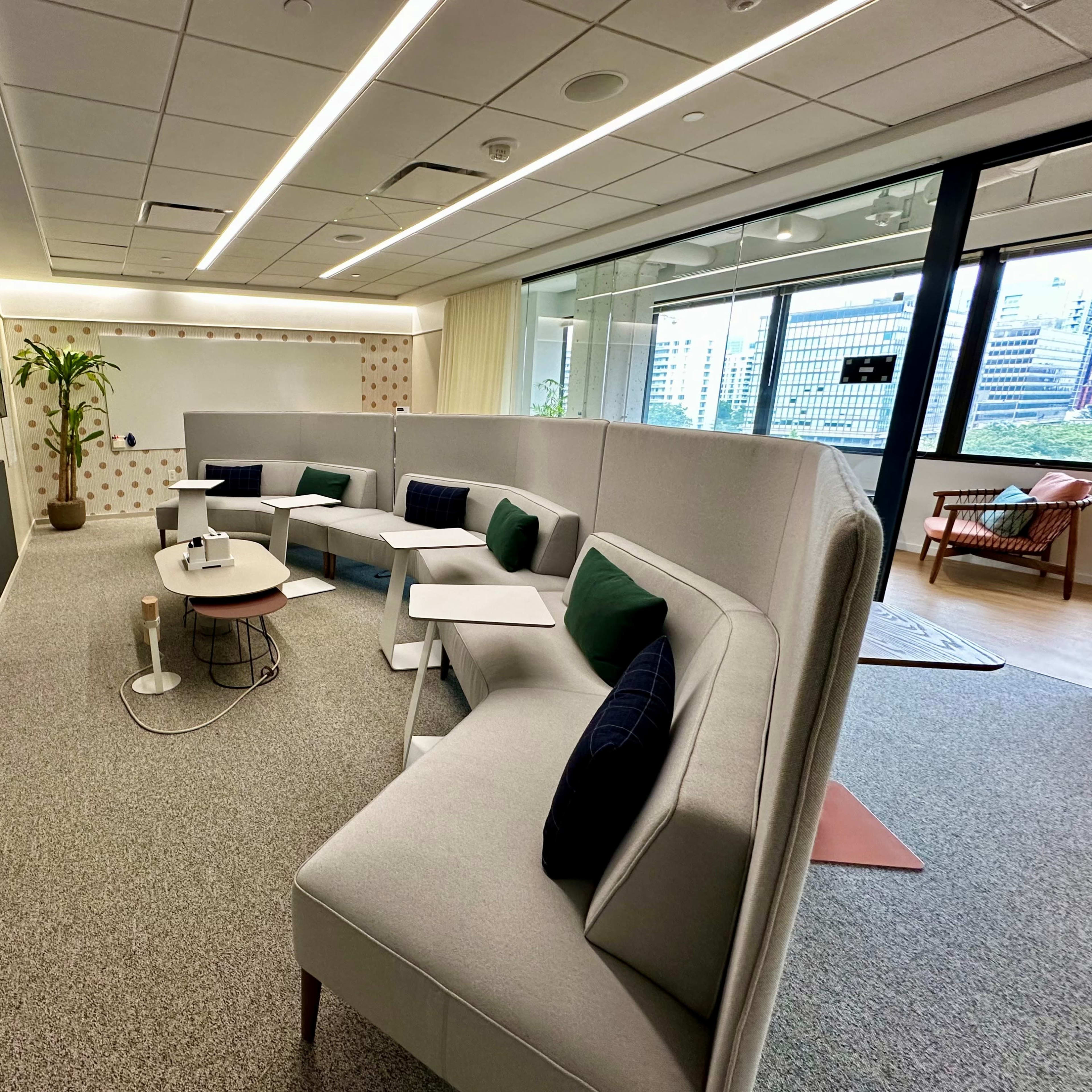 A modern office lounge area with modular seating, a television mounted on the wall, and large windows overlooking a cityscape.