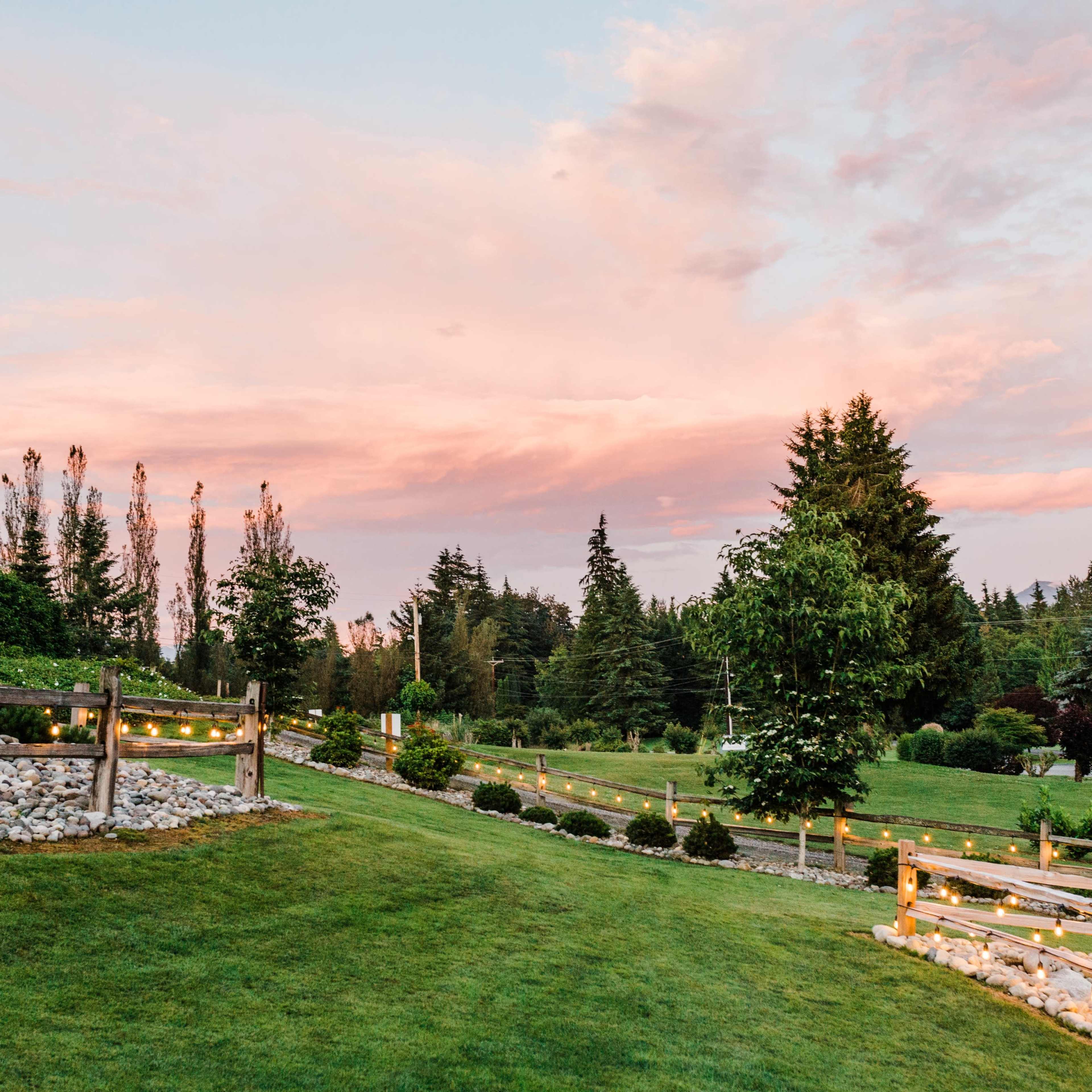 The scene depicts a landscaped area with rolling green grass, wooden fences, and trees under a pink and blue sky at sunset.