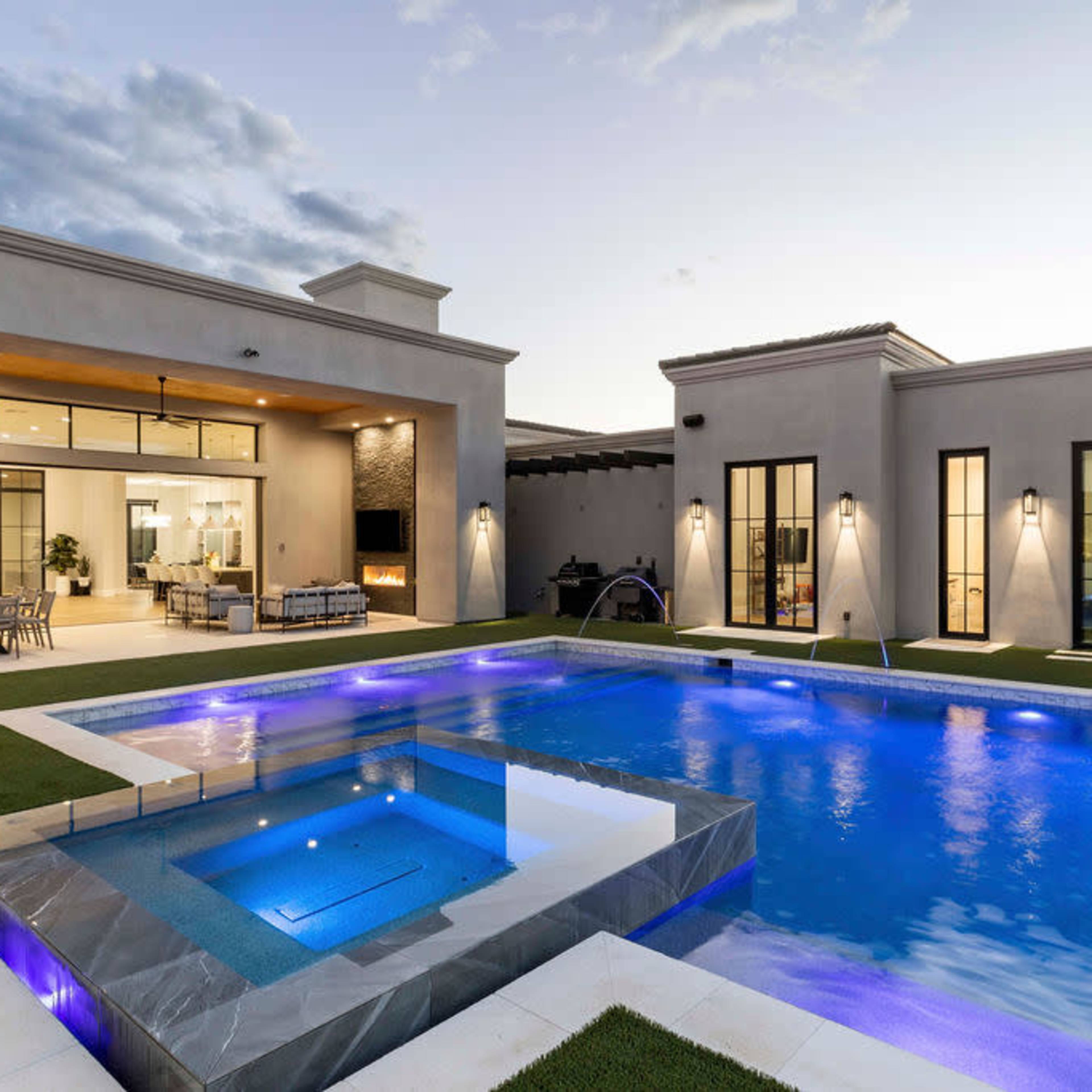 A modern house with a swimming pool featuring blue lighting, surrounded by green grass and outdoor seating areas at dusk.