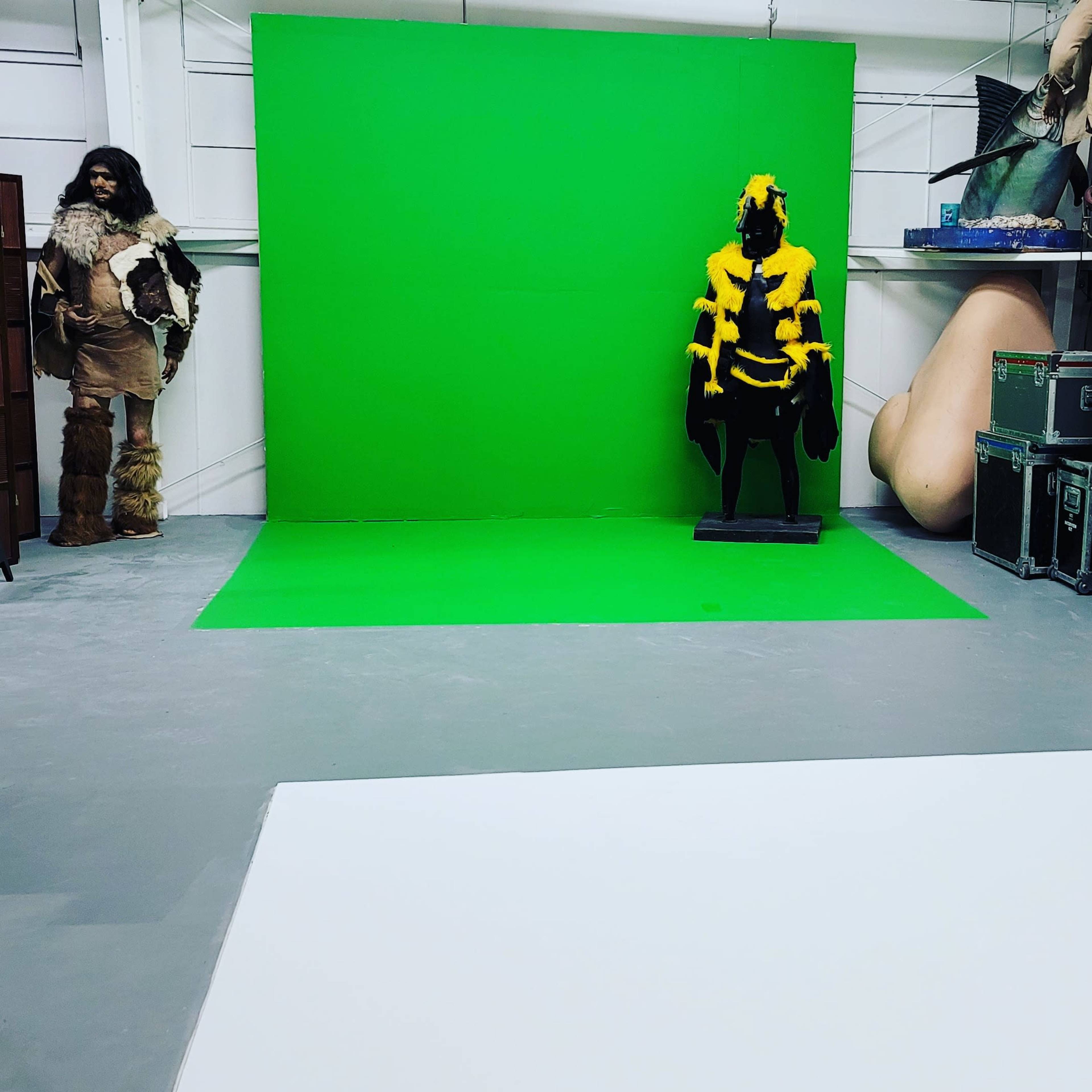 A studio space with a green screen backdrop, a white area on the floor, and two figures, one dressed in fur and the other in a yellow outfit, positioned nearby.