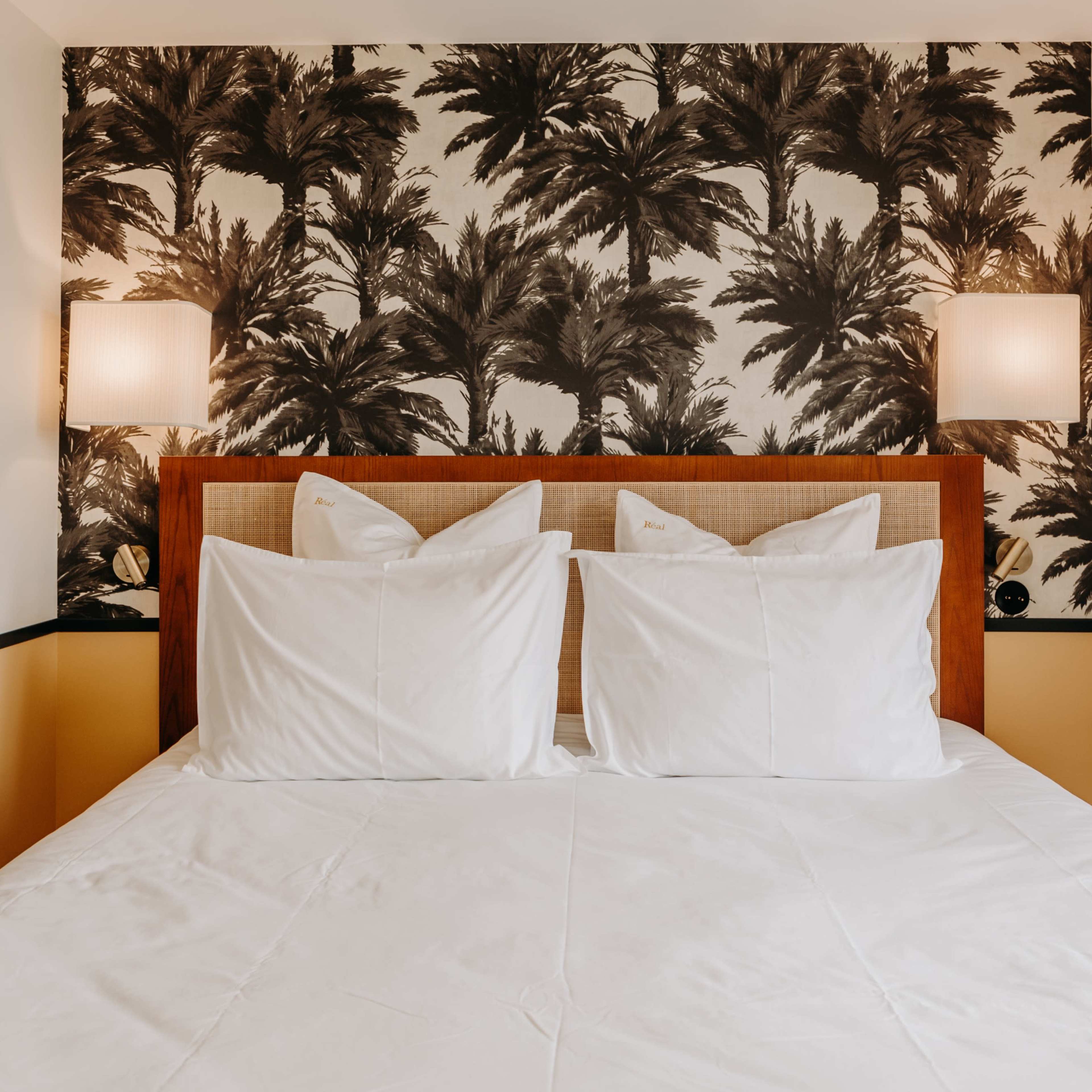 A bedroom with a bed featuring white pillows against a backdrop of palm-patterned wallpaper.