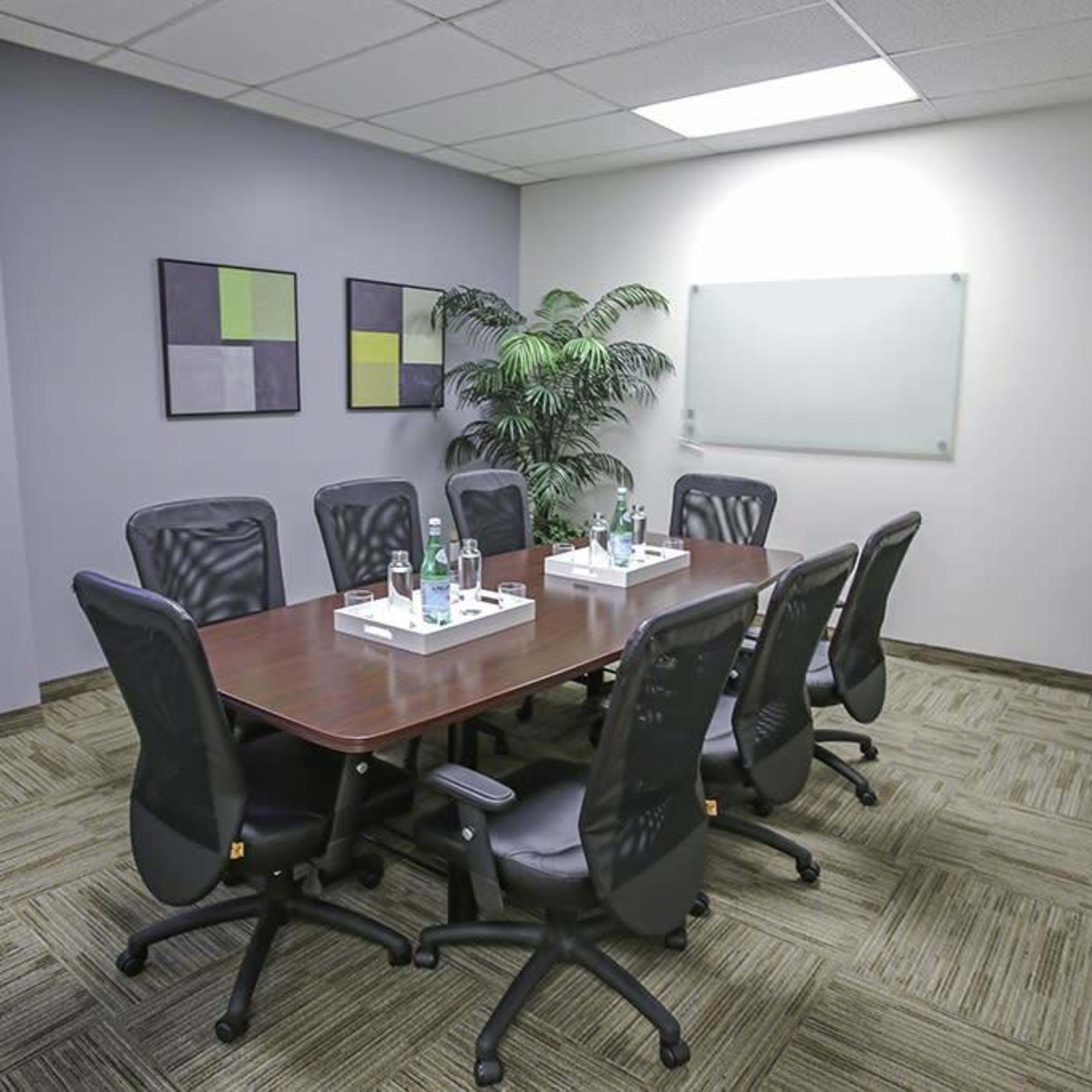 A conference room features a large table surrounded by eight chairs, with a plant in the corner and minimal artwork on the walls.