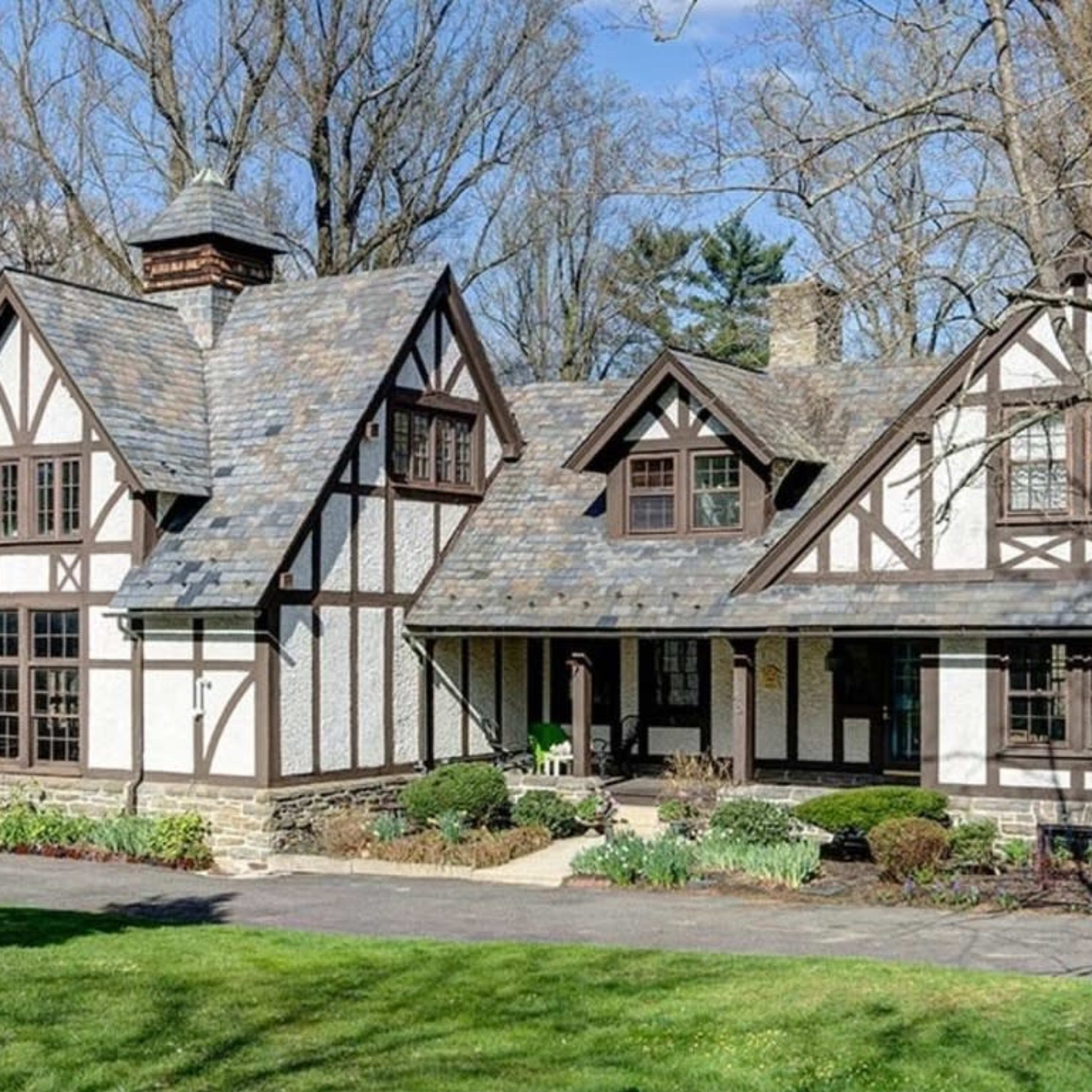 A large, two-story house with a steeply pitched roof, exposed wooden beams, and a stone foundation, surrounded by greenery.