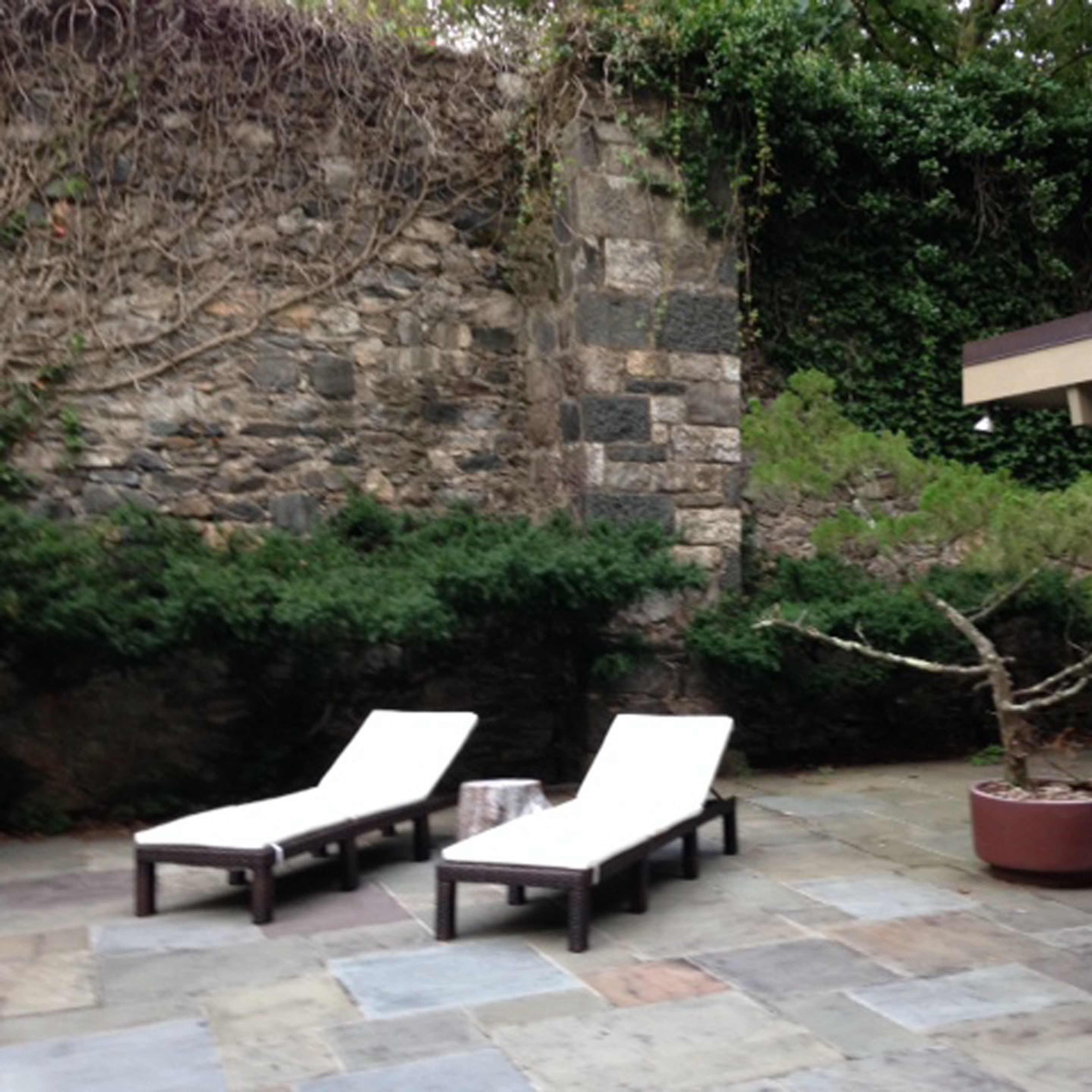 A patio with two white lounge chairs beside a stone wall and a potted tree.