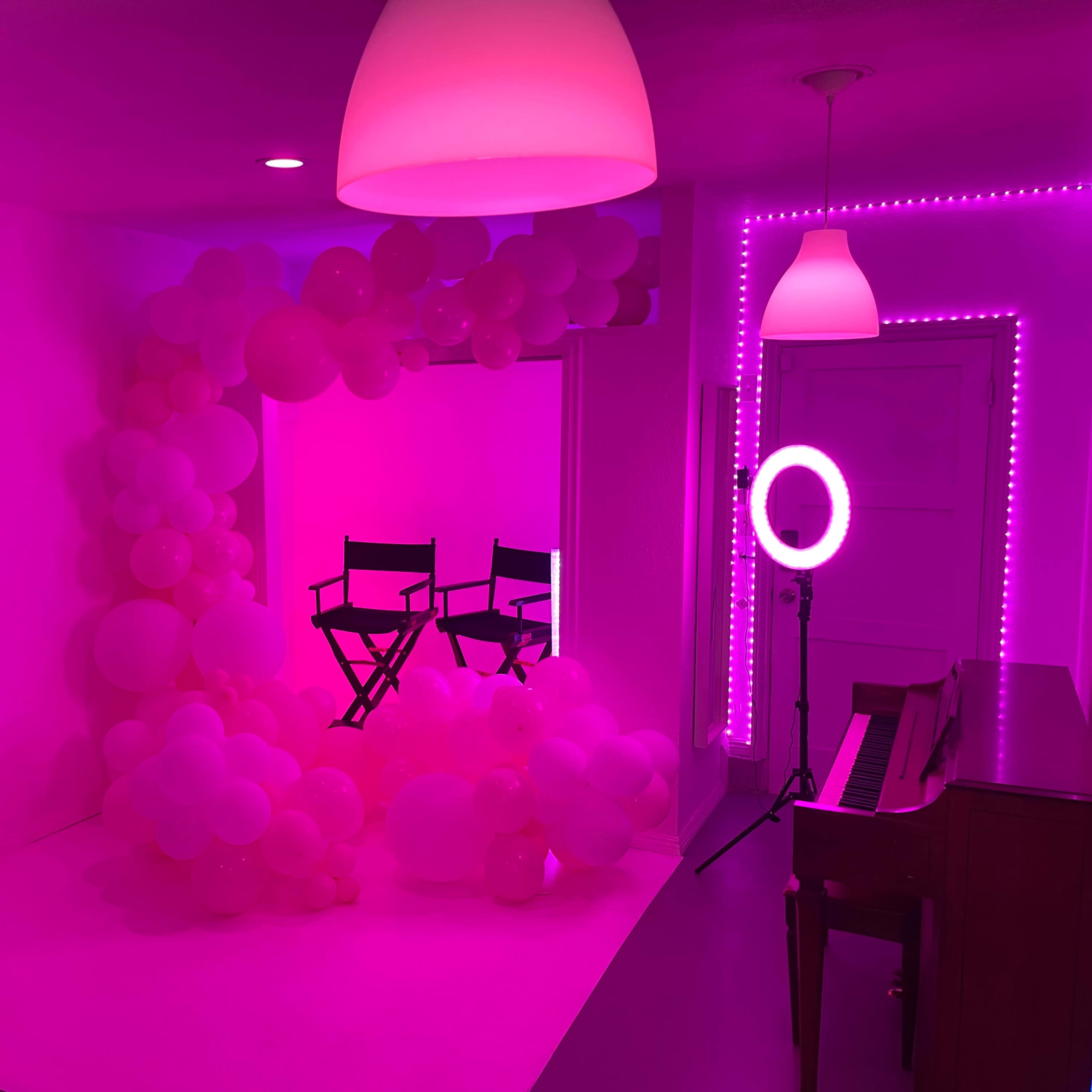 A brightly lit room decorated with pink balloons, featuring two director's chairs, a ring light, and a piano.