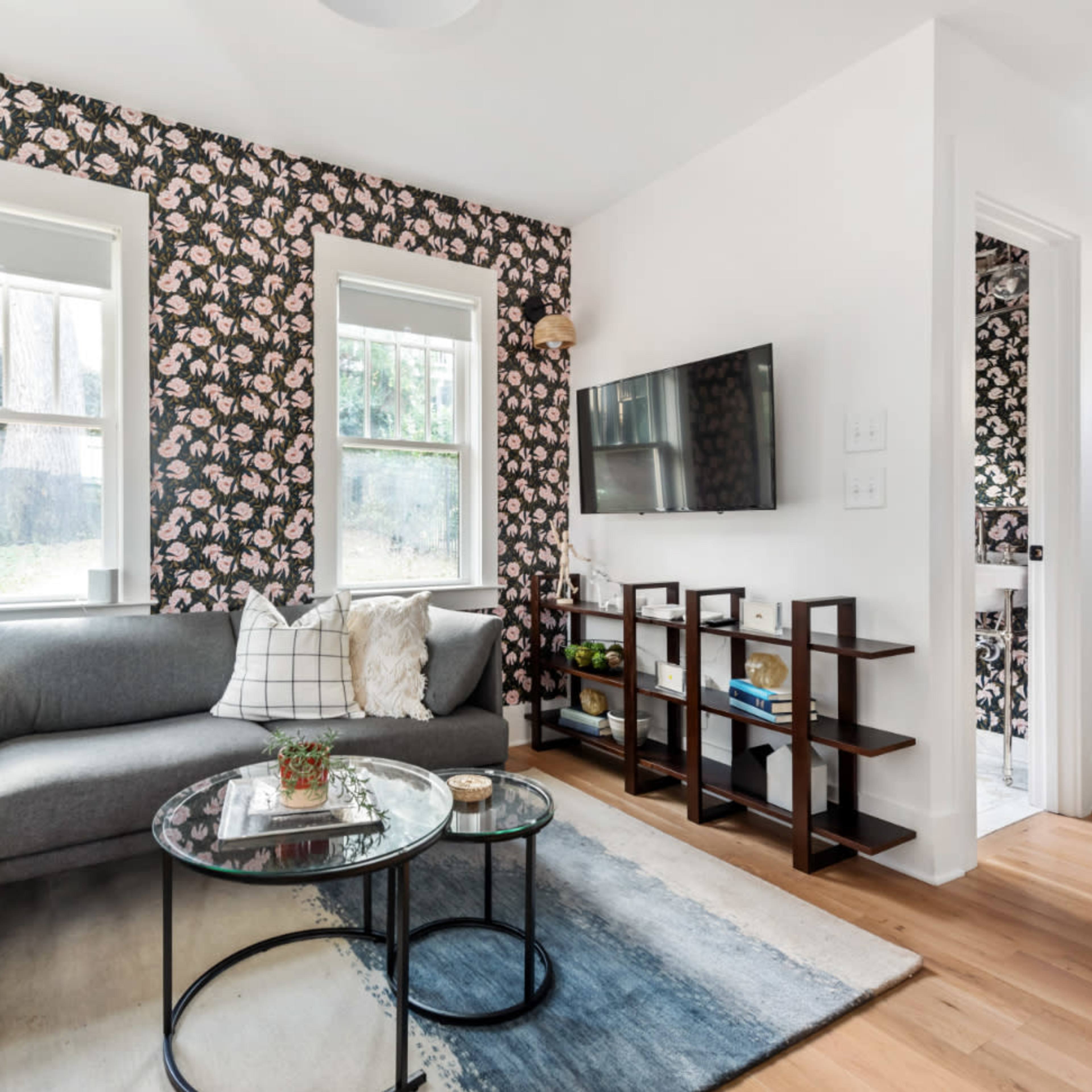 A modern living room featuring a gray sectional sofa, a glass coffee table, floral wallpaper, and a wall-mounted television.