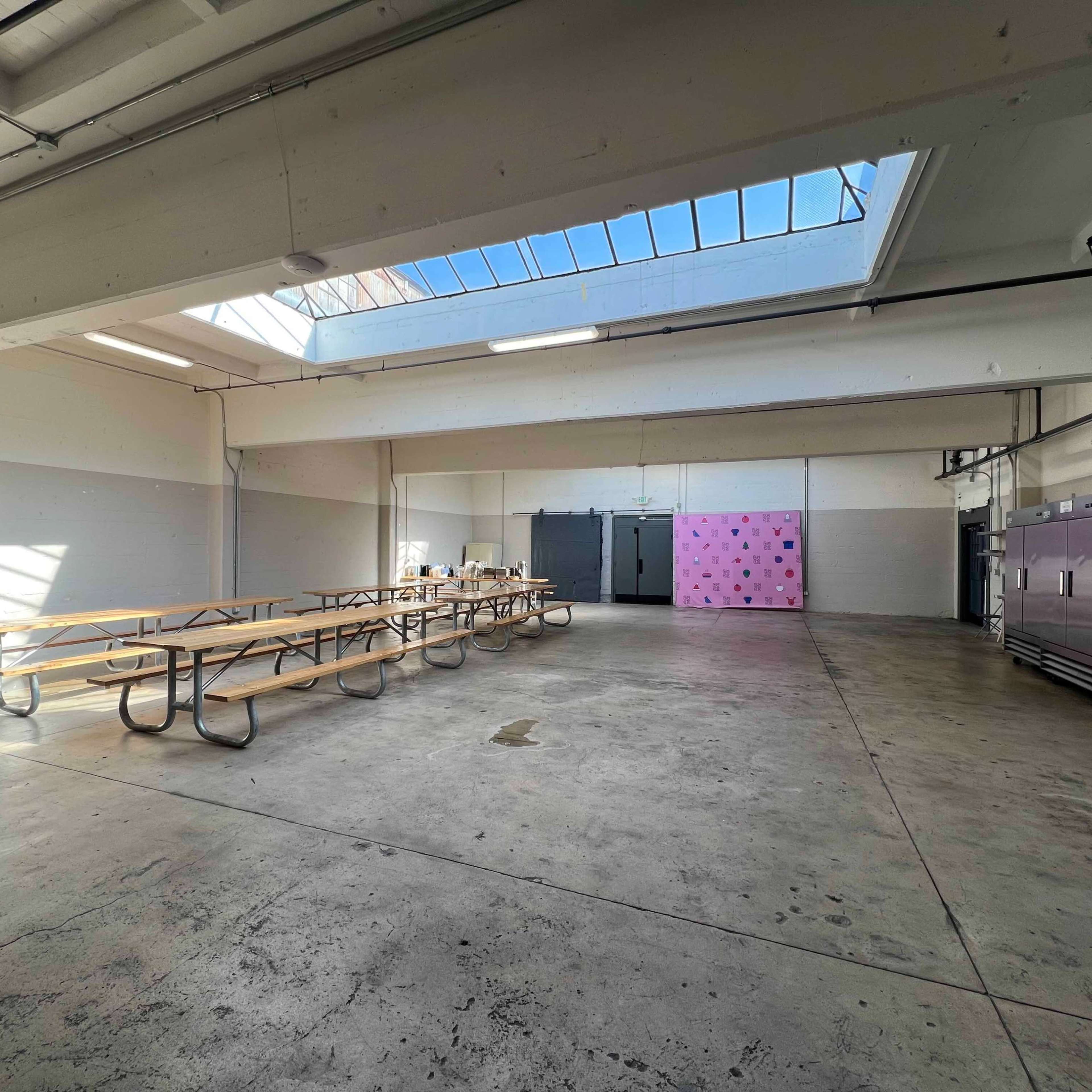 A spacious, empty room with rows of wooden picnic tables, a pink wall at one end, and large refrigerators along the side.