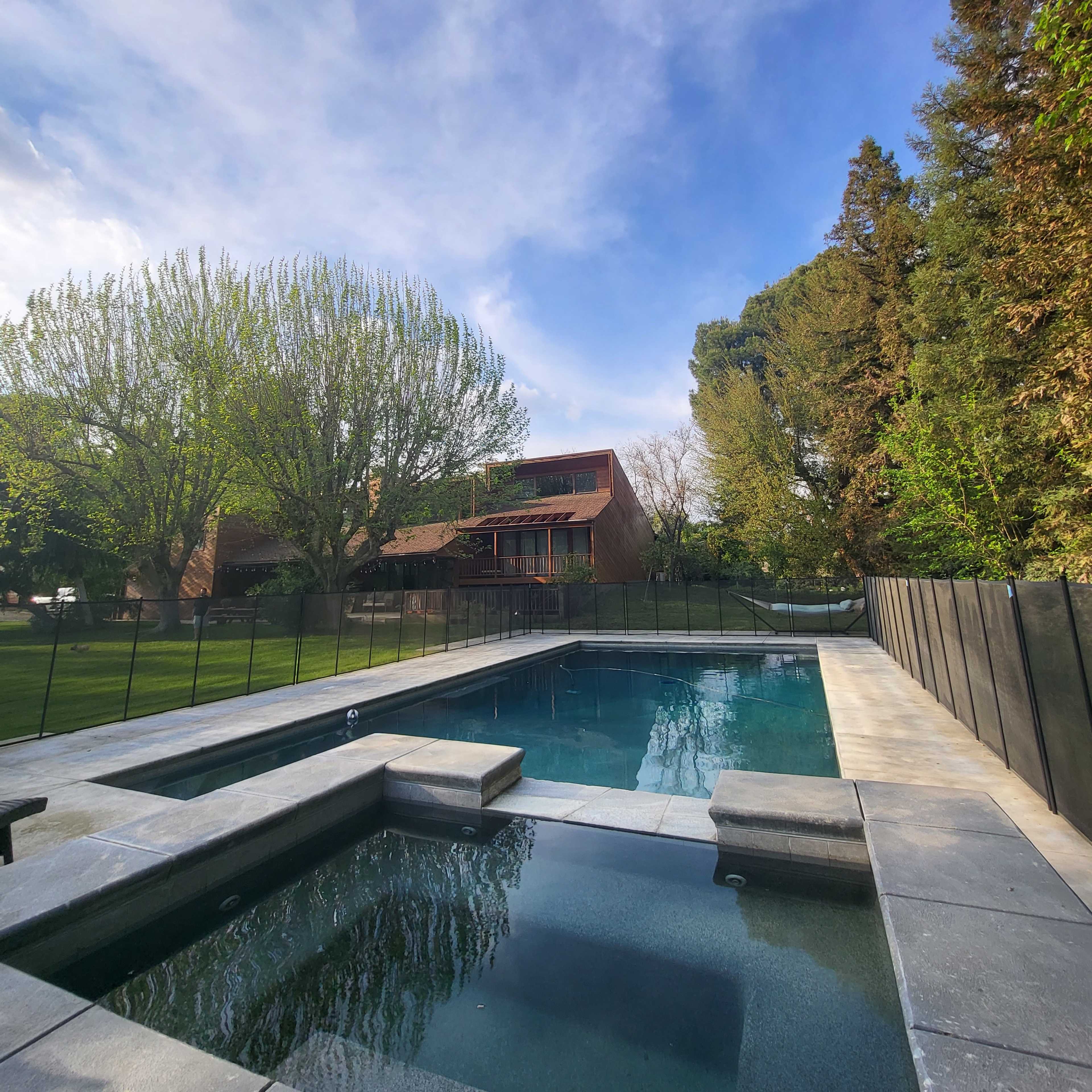 A rectangular pool with a connected spa, surrounded by a black fence, grassy areas, and trees, under a clear sky.