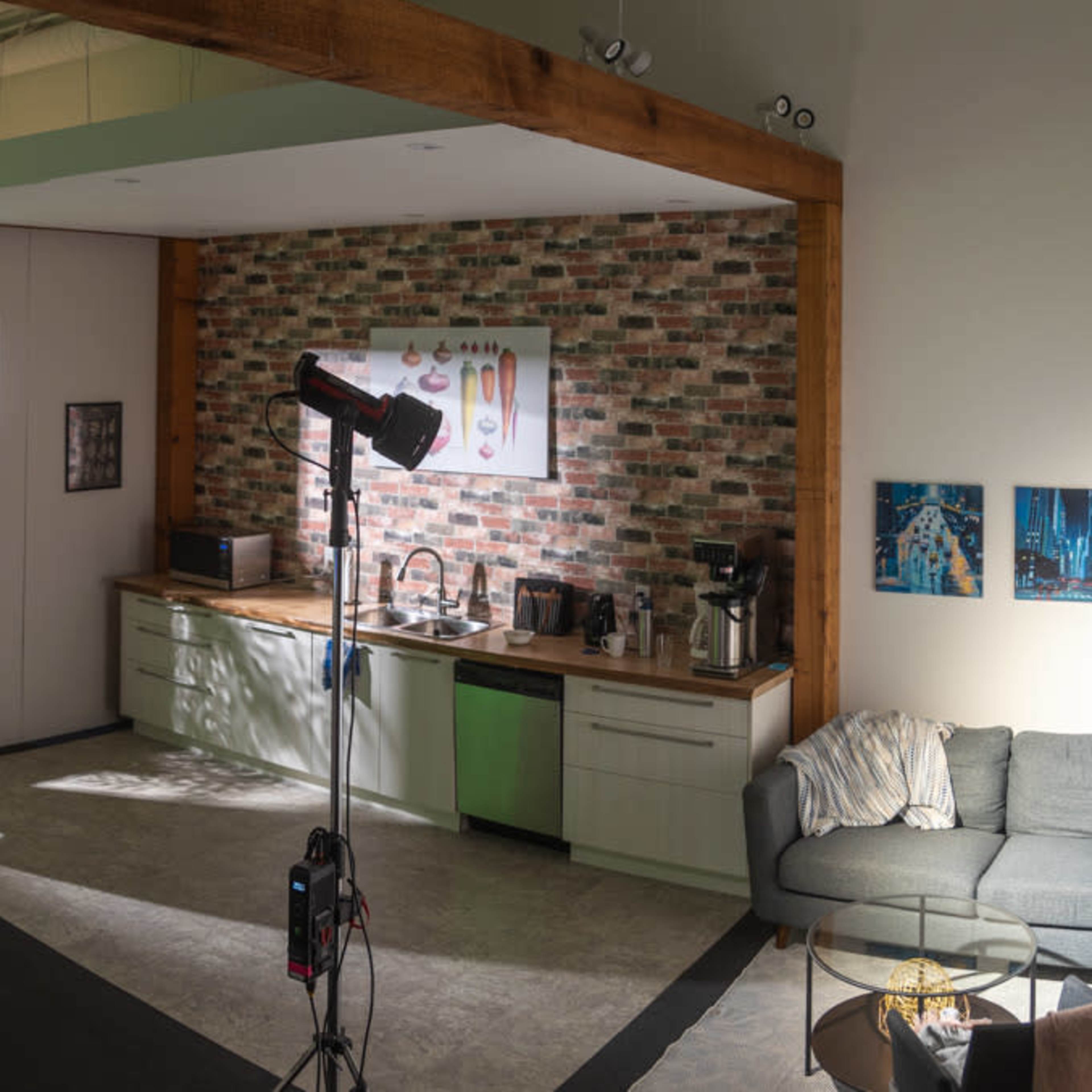 A modern indoor space featuring a kitchen with a brick backsplash, a seating area with a gray couch, and multiple studio lights set up for photography.