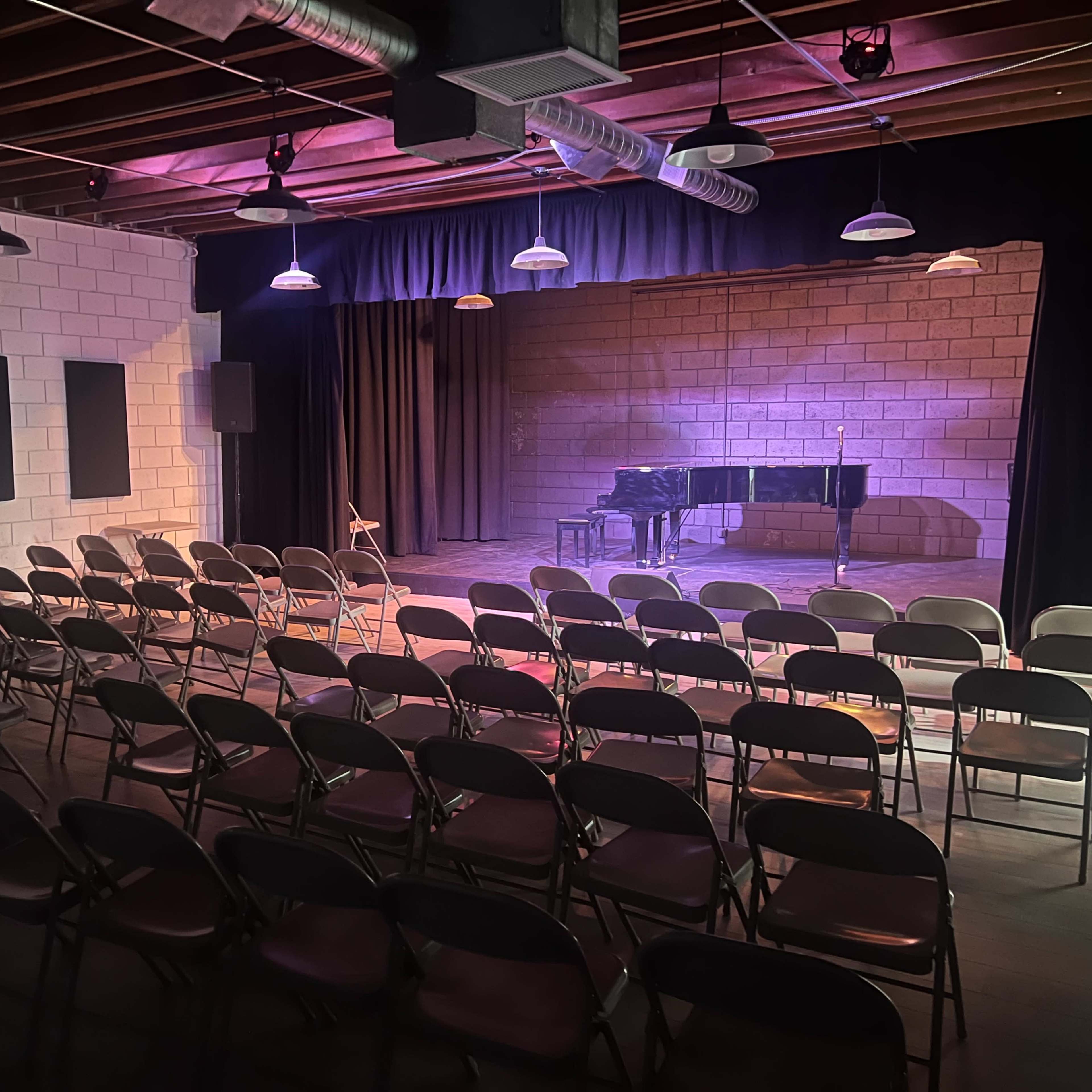 A performance space with empty rows of metal chairs facing a stage that features a grand piano and soft lighting.