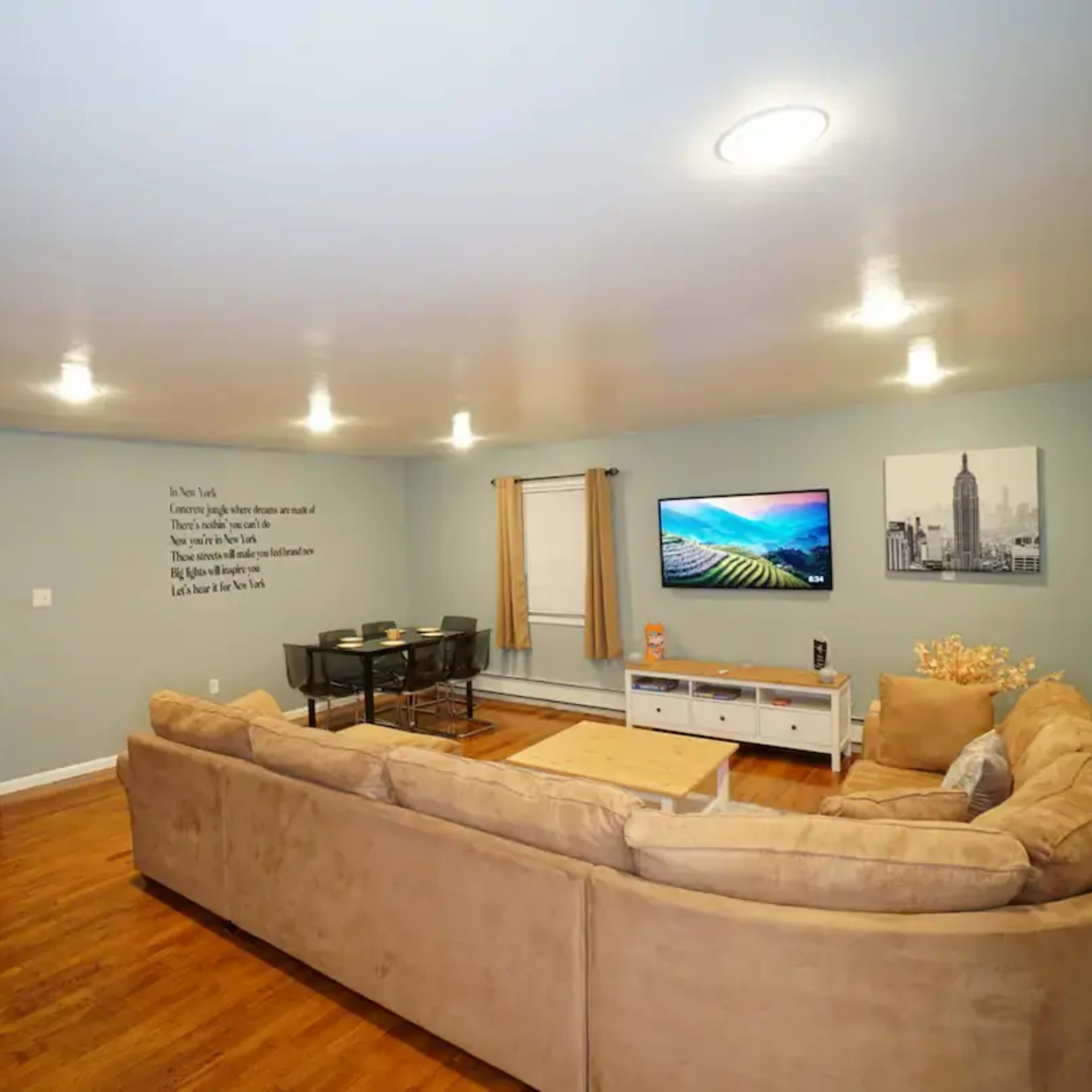 A spacious living room with a large beige sectional sofa, a wall-mounted television, and a dining table set against a blue wall with decorative elements.