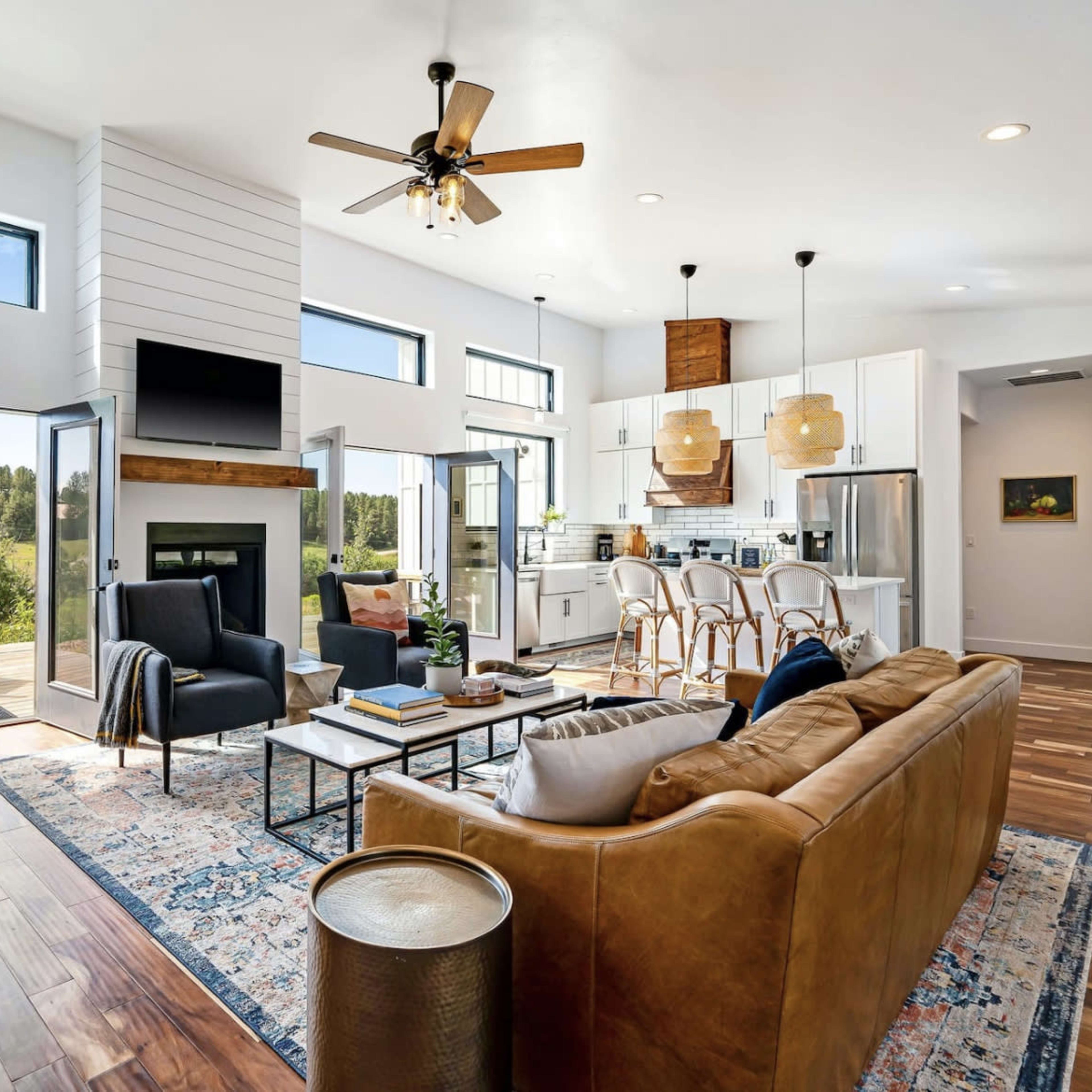 A modern living room with large windows and an open floor plan that connects to a kitchen area, featuring a brown leather sofa, wooden flooring, and a view of greenery outside.
