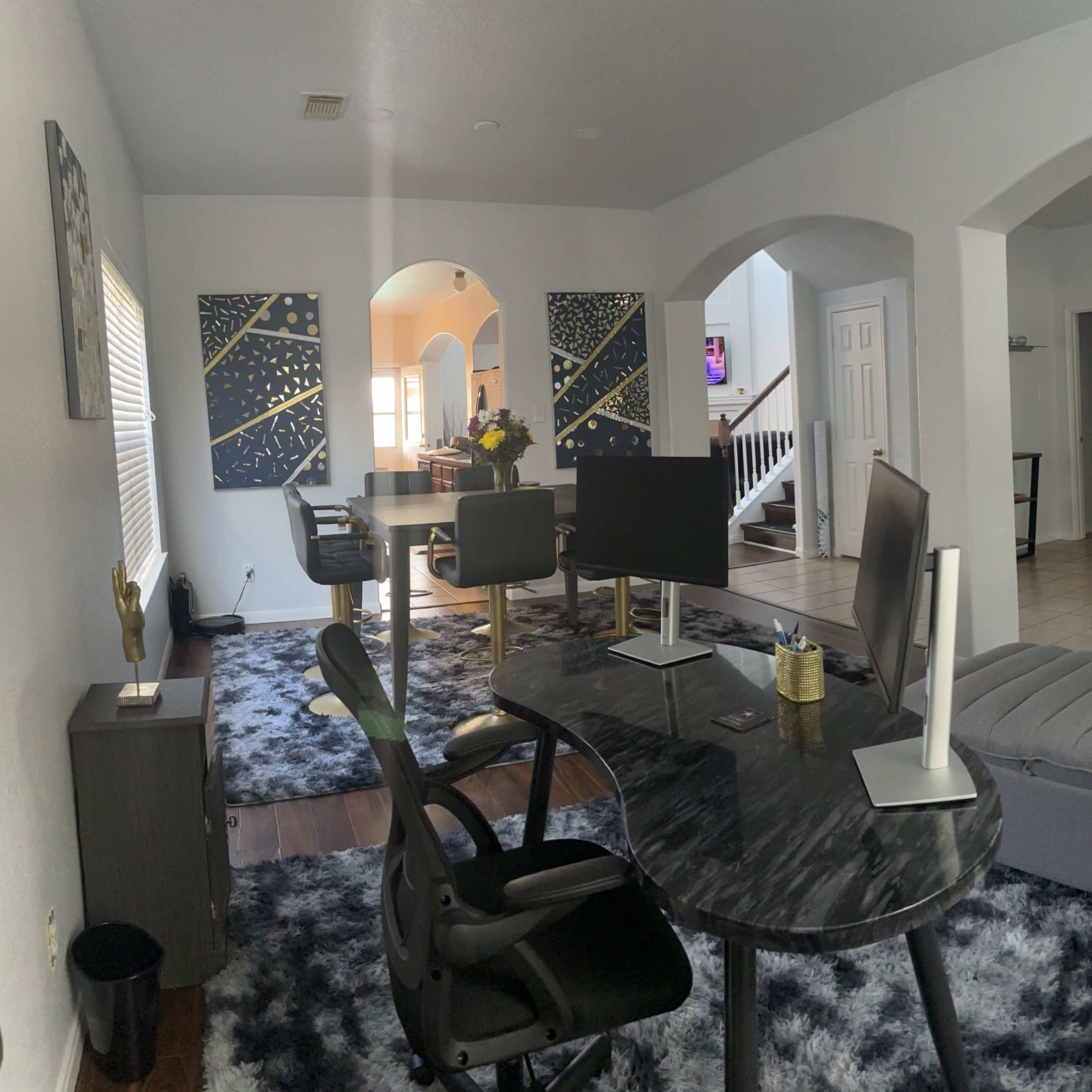 A spacious living area featuring a dark marble desk with computer monitors, a gray ottoman, and artistic wall decorations, alongside an open archway leading to another room.