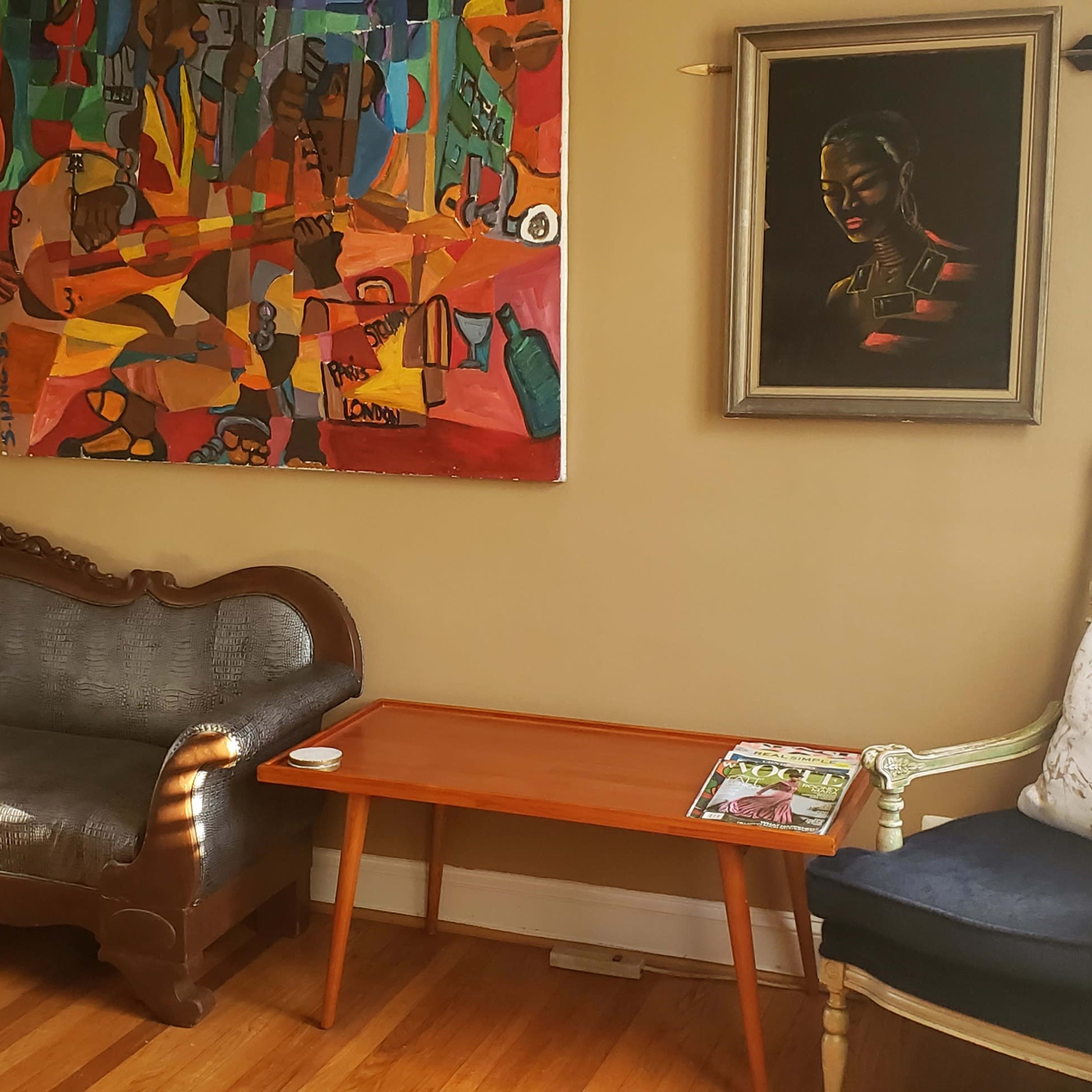 A living room featuring a dark sofa, an orange abstract painting on the wall, a wooden side table with a magazine, and a decorative chair with a cushion.