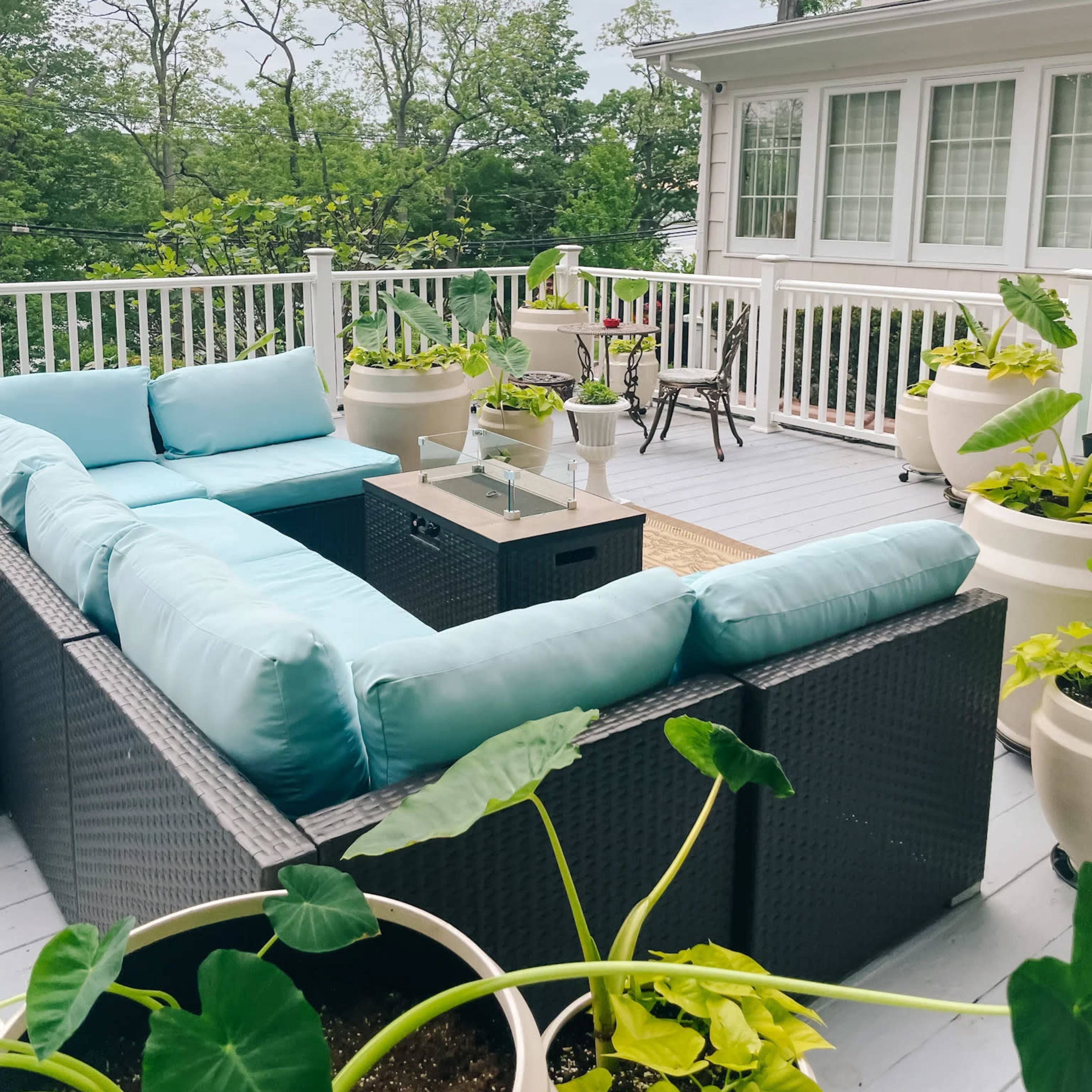 A spacious outdoor patio features a light blue sectional sofa, a small table, and various potted plants, surrounded by trees and a house in the background.