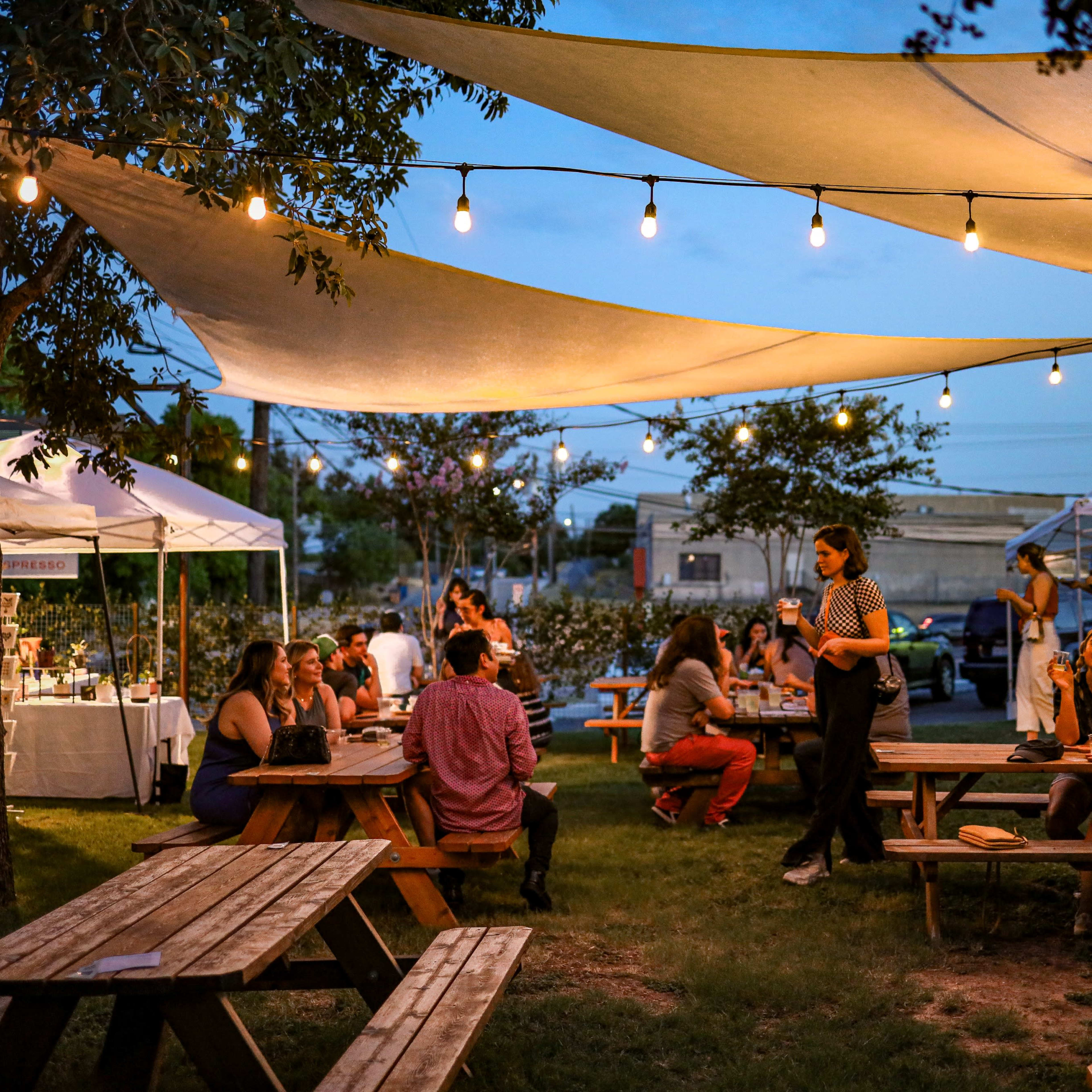 A lively outdoor market features people socializing at wooden picnic tables under string lights and canopies at dusk.