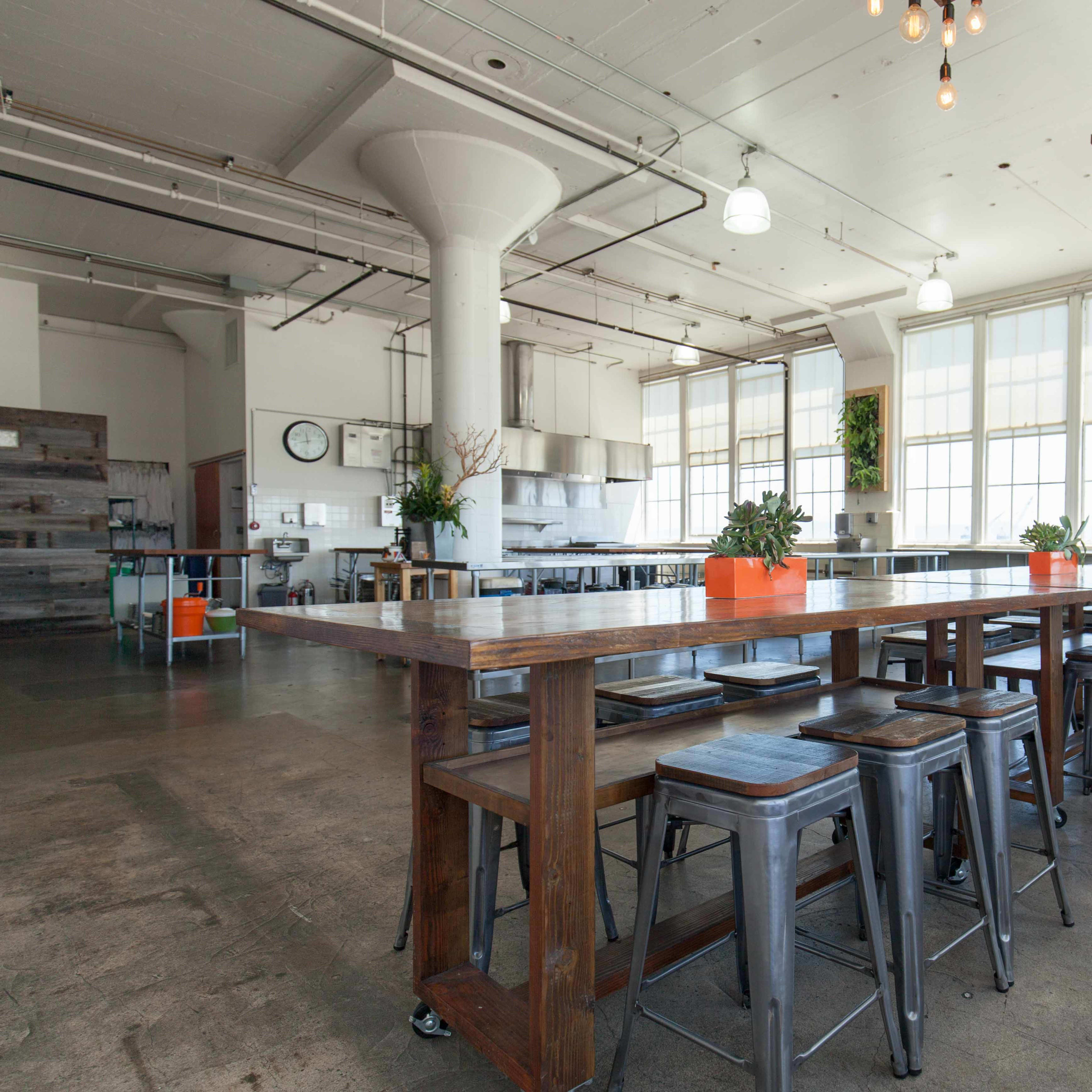 A spacious, modern dining area with long wooden tables and metal stools, surrounded by large windows and hanging plants.