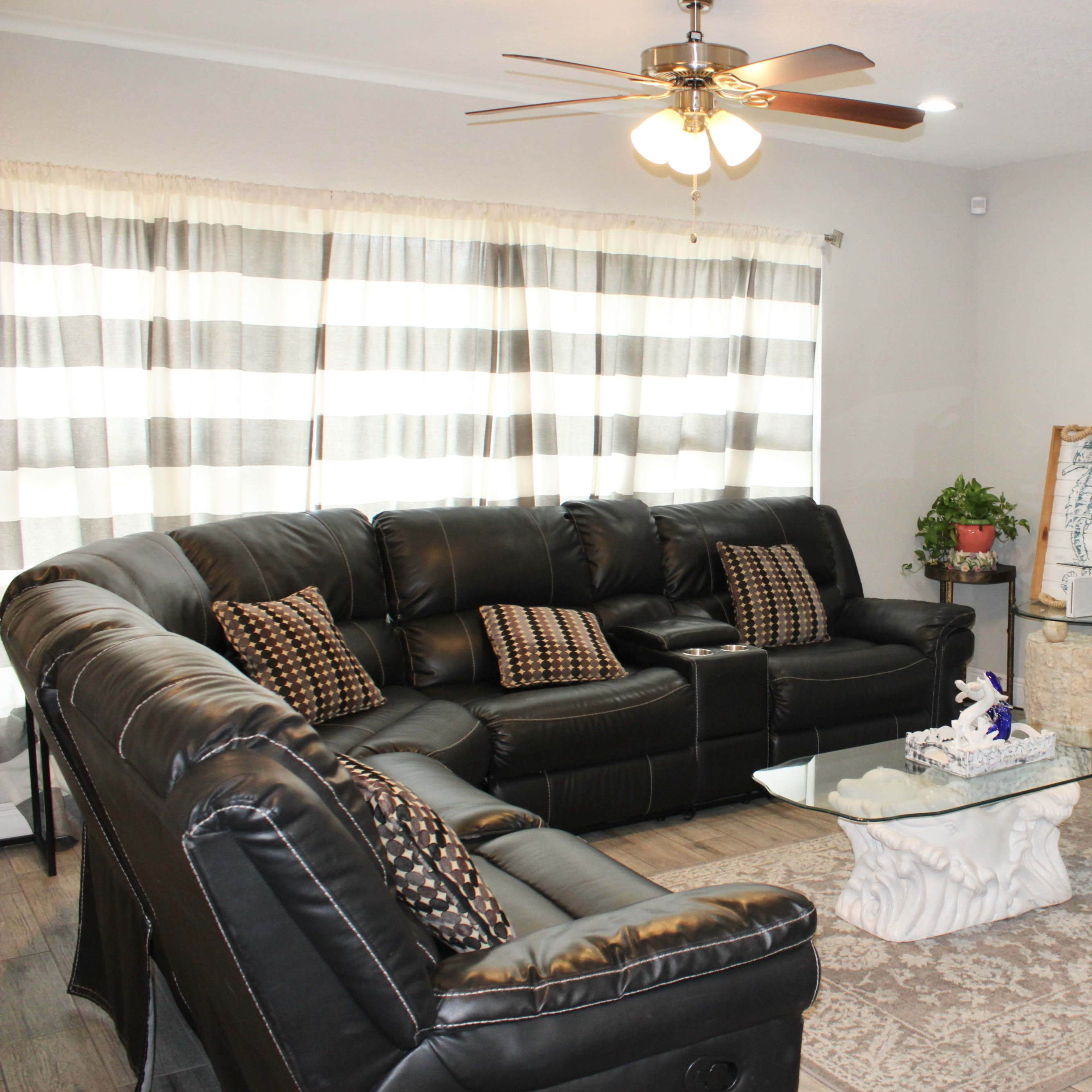 A living room with a black leather sectional sofa, a glass coffee table on a patterned rug, and a wall-mounted television.