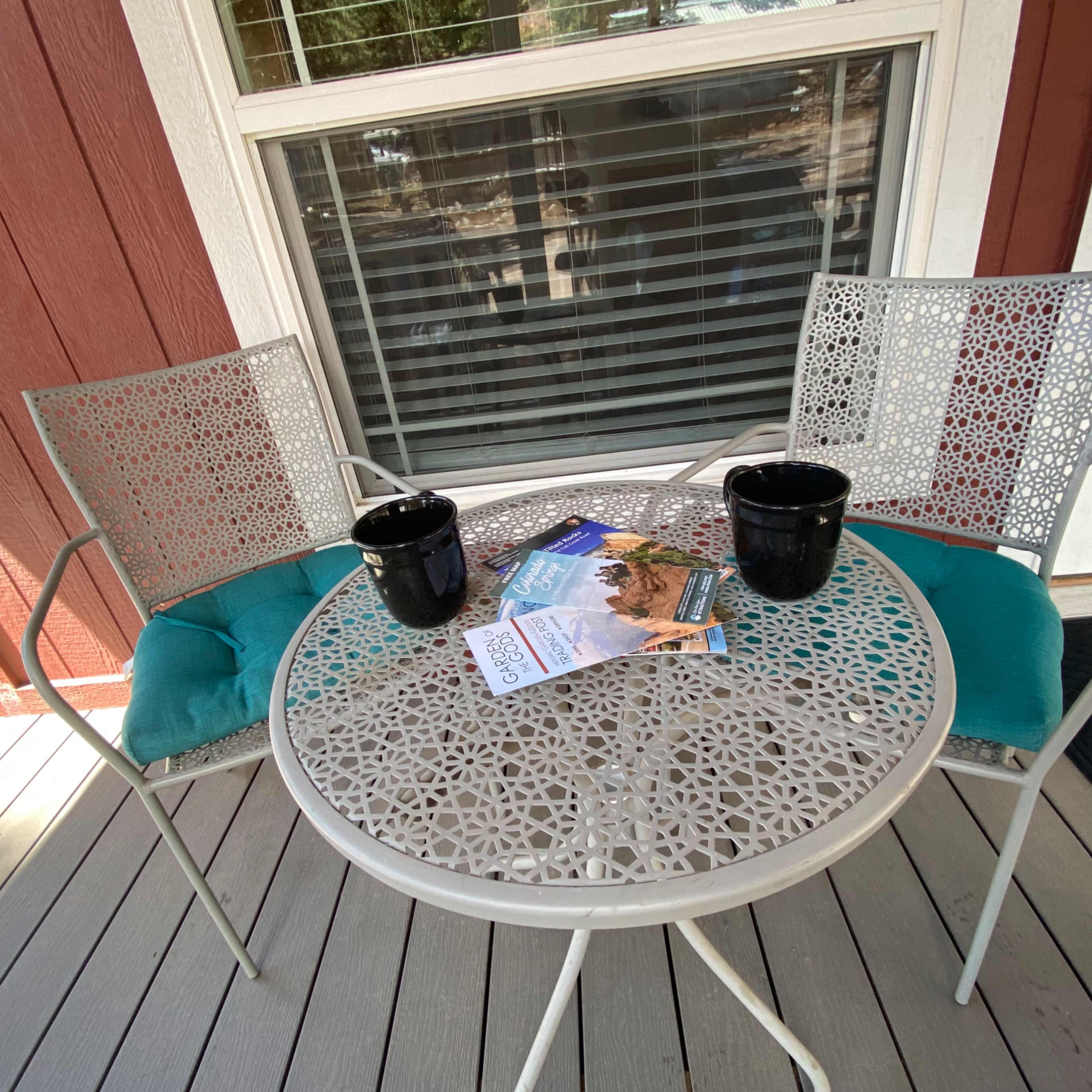 A small round table with two metal chairs and black planters sits on a wooden deck beside a door.