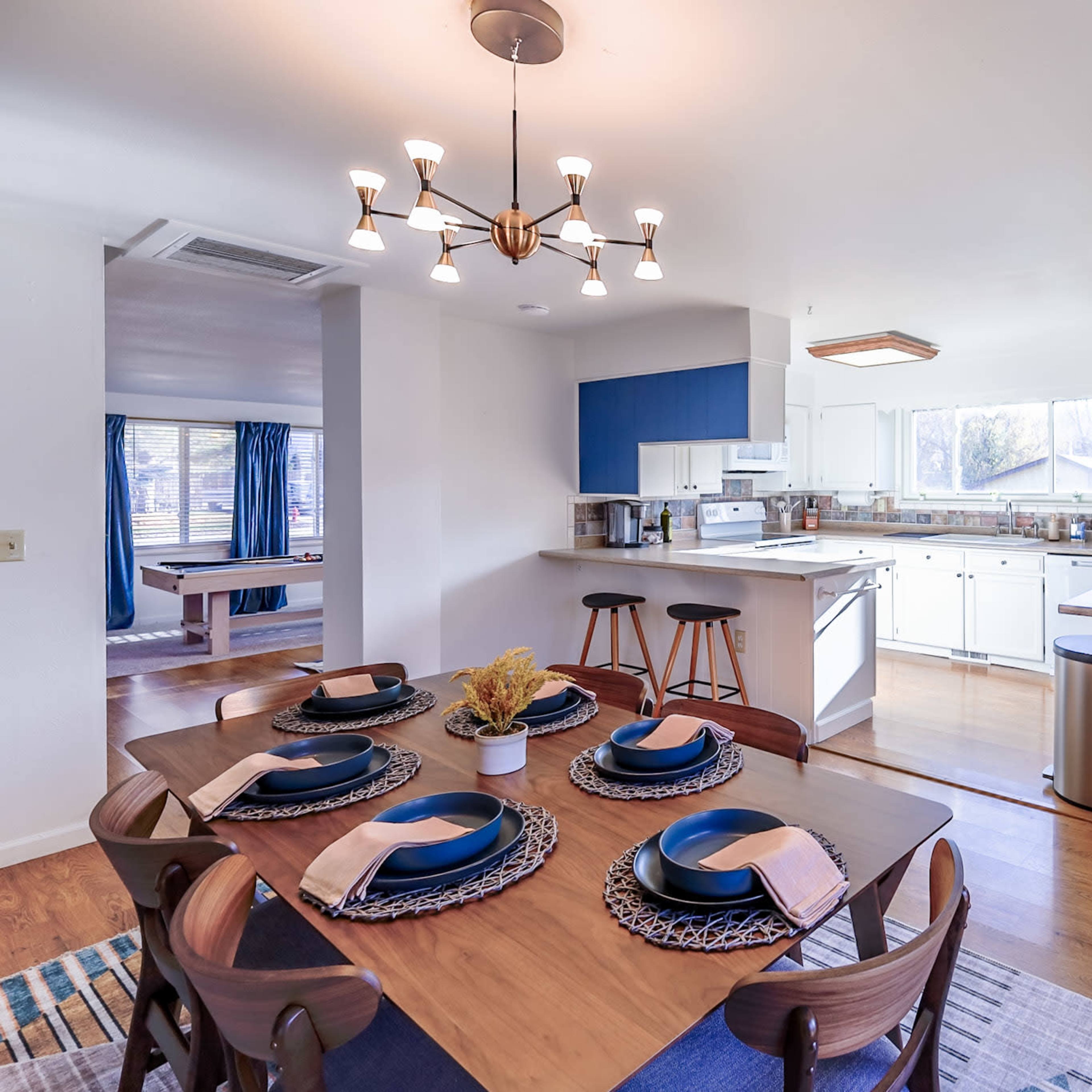 A modern dining area features a table set for six, adjacent to an open kitchen with blue cabinets and stainless steel appliances.