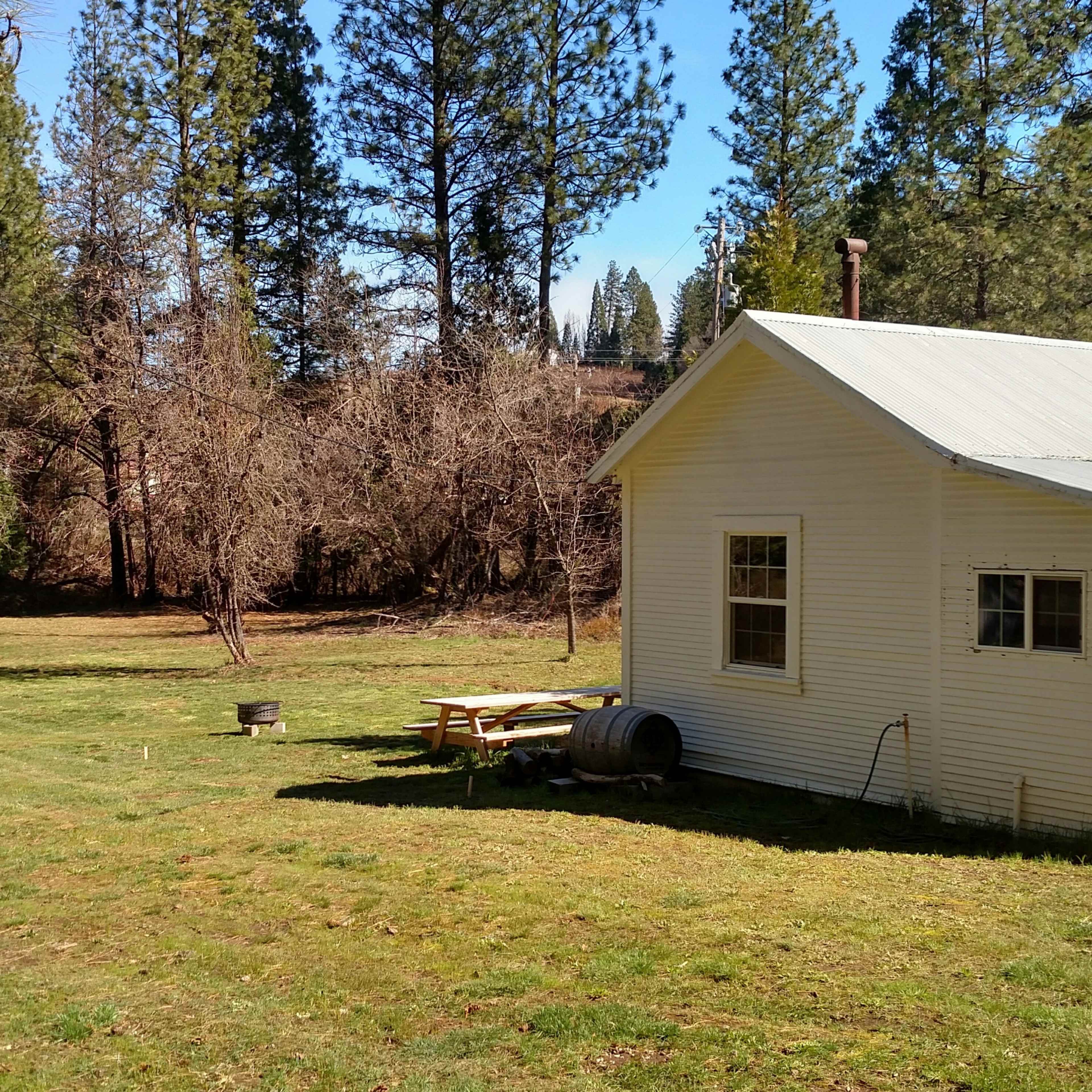 A small white cottage sits on a grassy area surrounded by tall trees and an open field.
