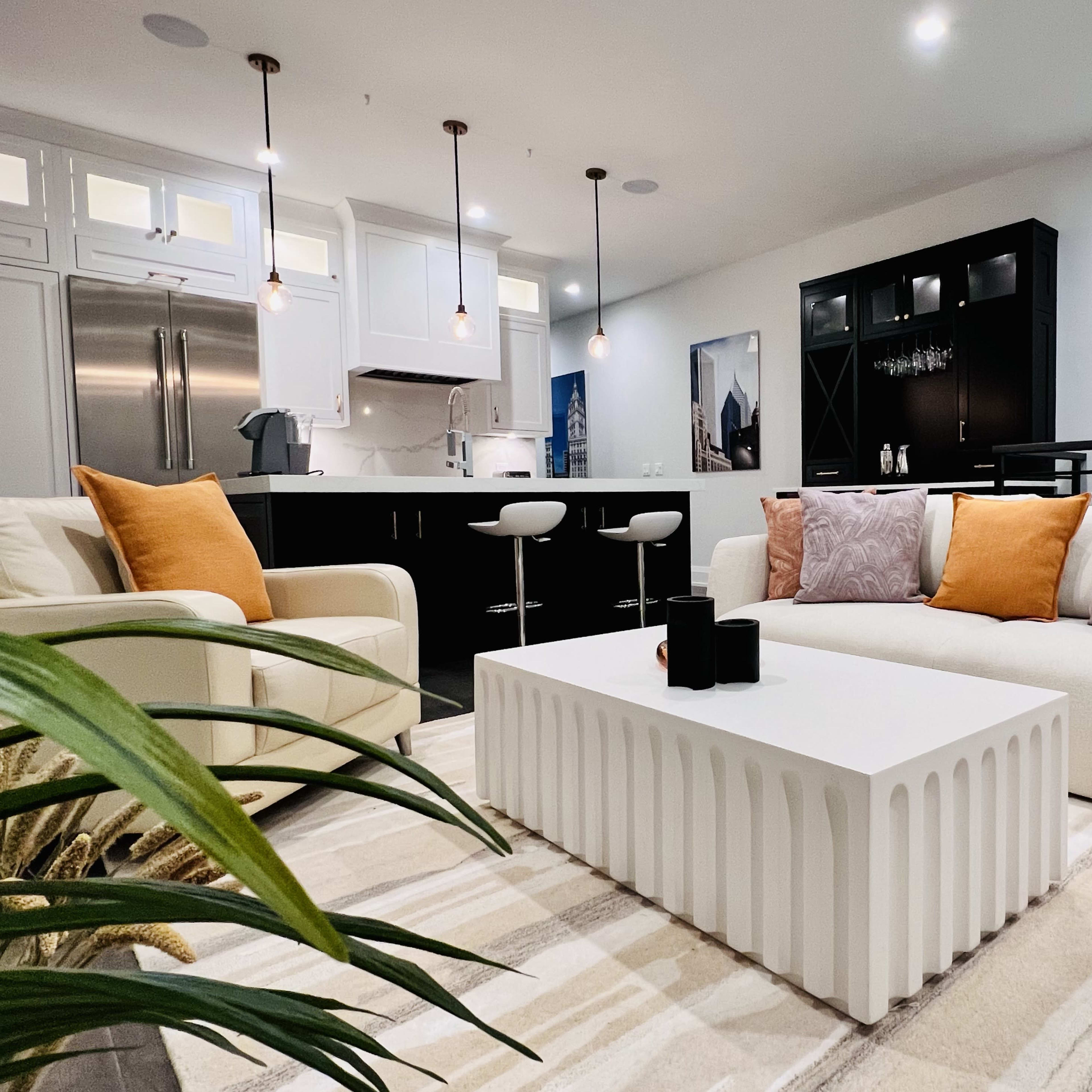 A modern living room with a white sofa and armchair, a minimalist coffee table, and a kitchen area in the background.