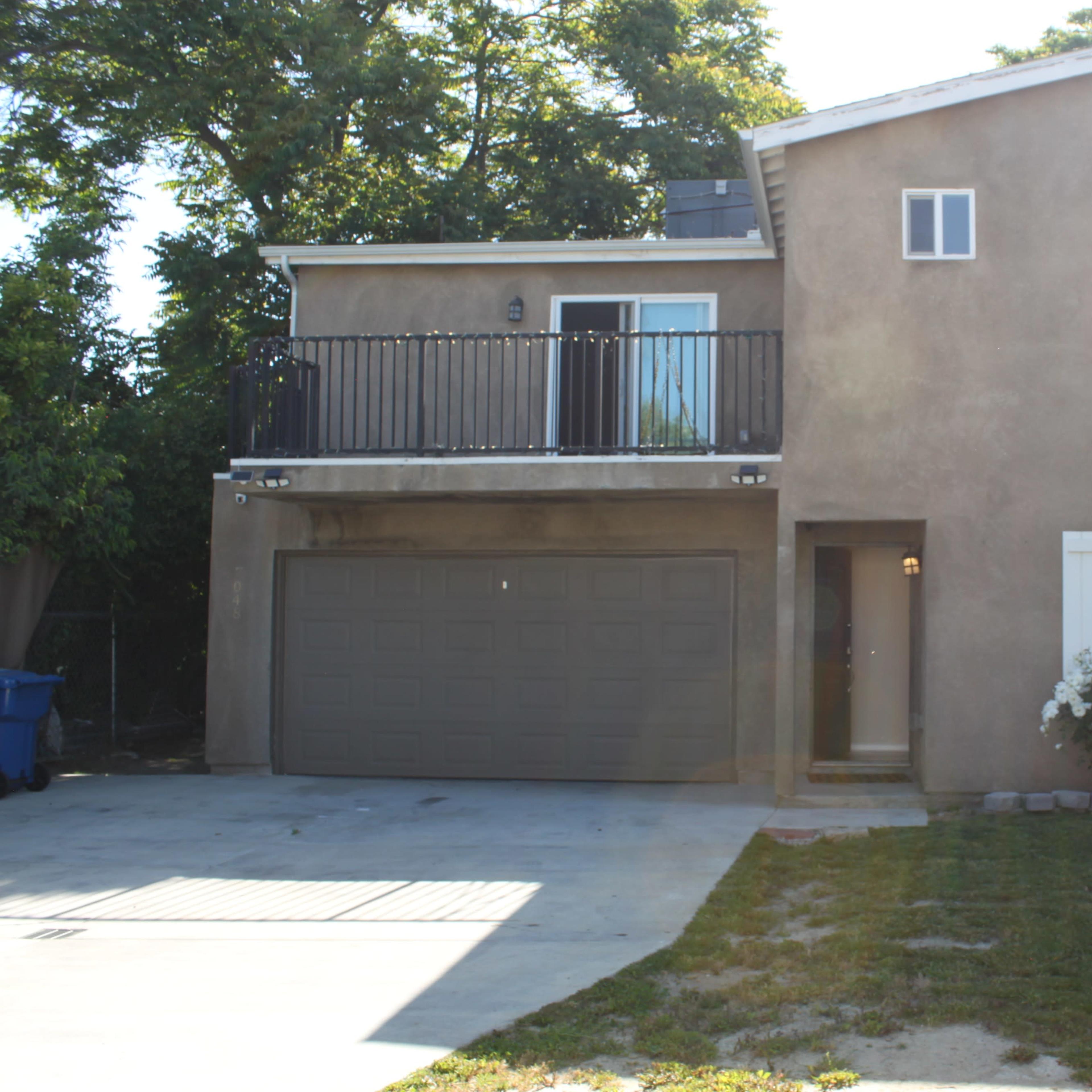 A two-story house features a balcony above a two-car garage, with a front yard and a paved driveway.