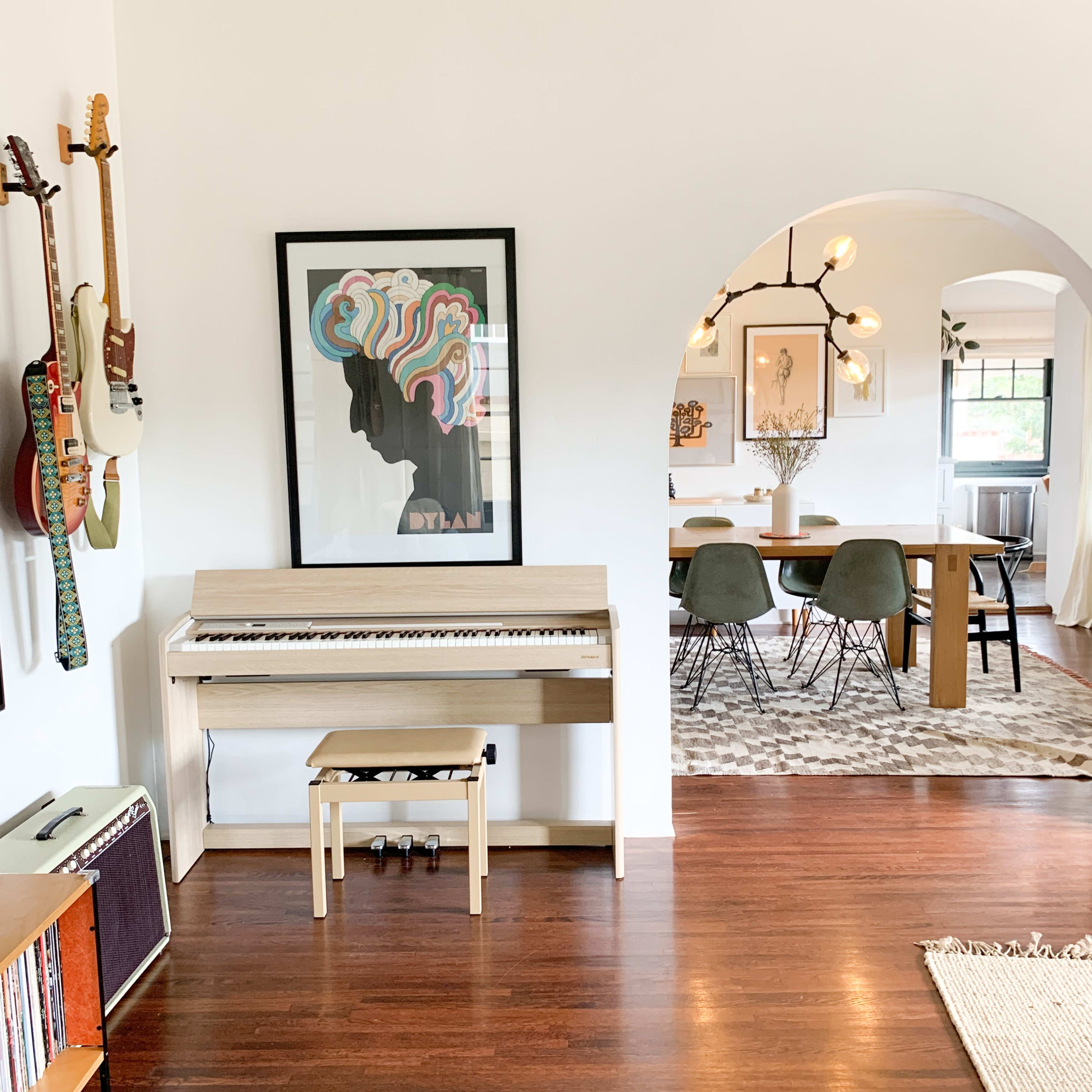 A living space featuring a digital piano under a colorful framed print, with guitars mounted on the wall and a dining area visible through an archway.