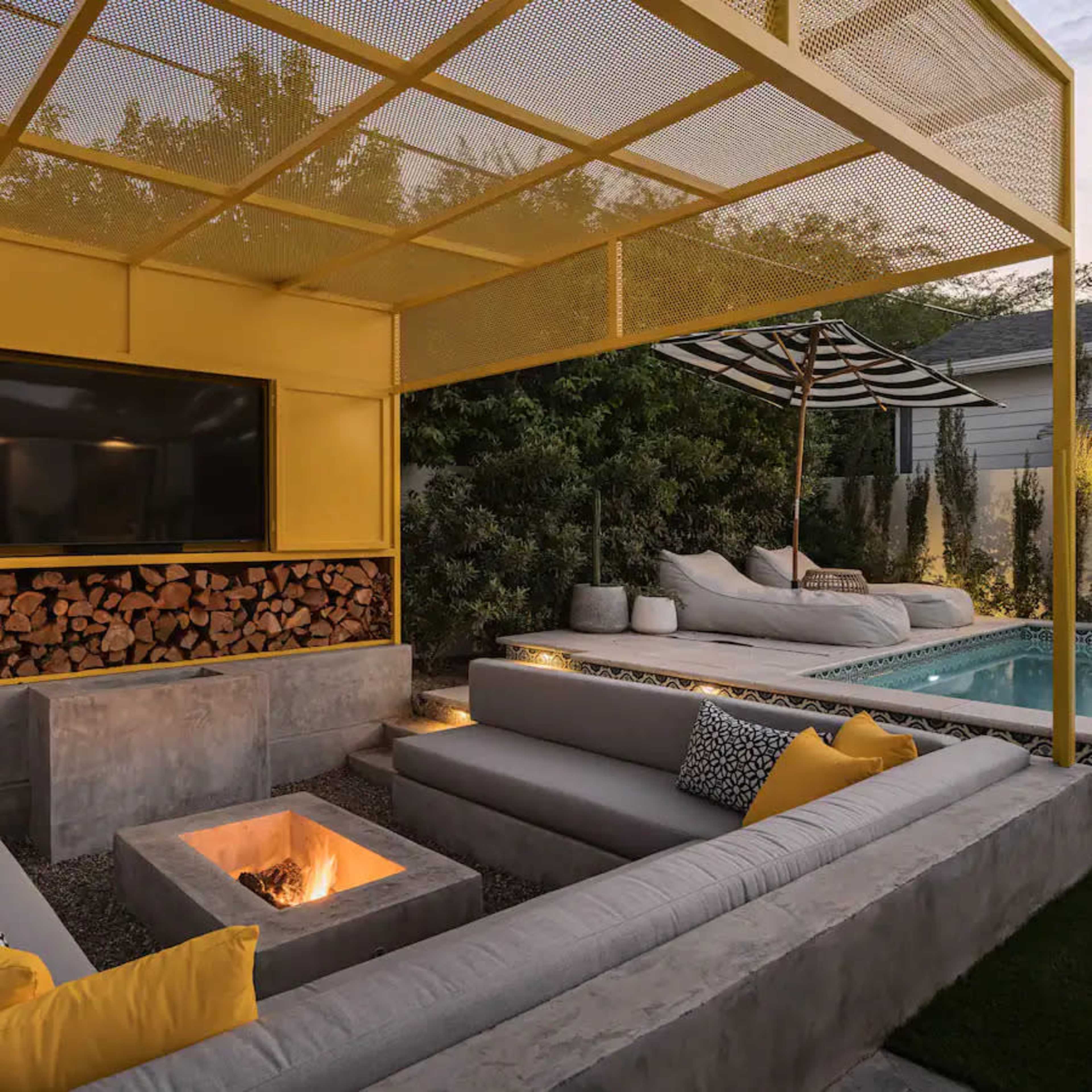 A modern outdoor lounge area with a fire pit, gray seating, a television on a yellow-framed pergola, and a pool in the background.